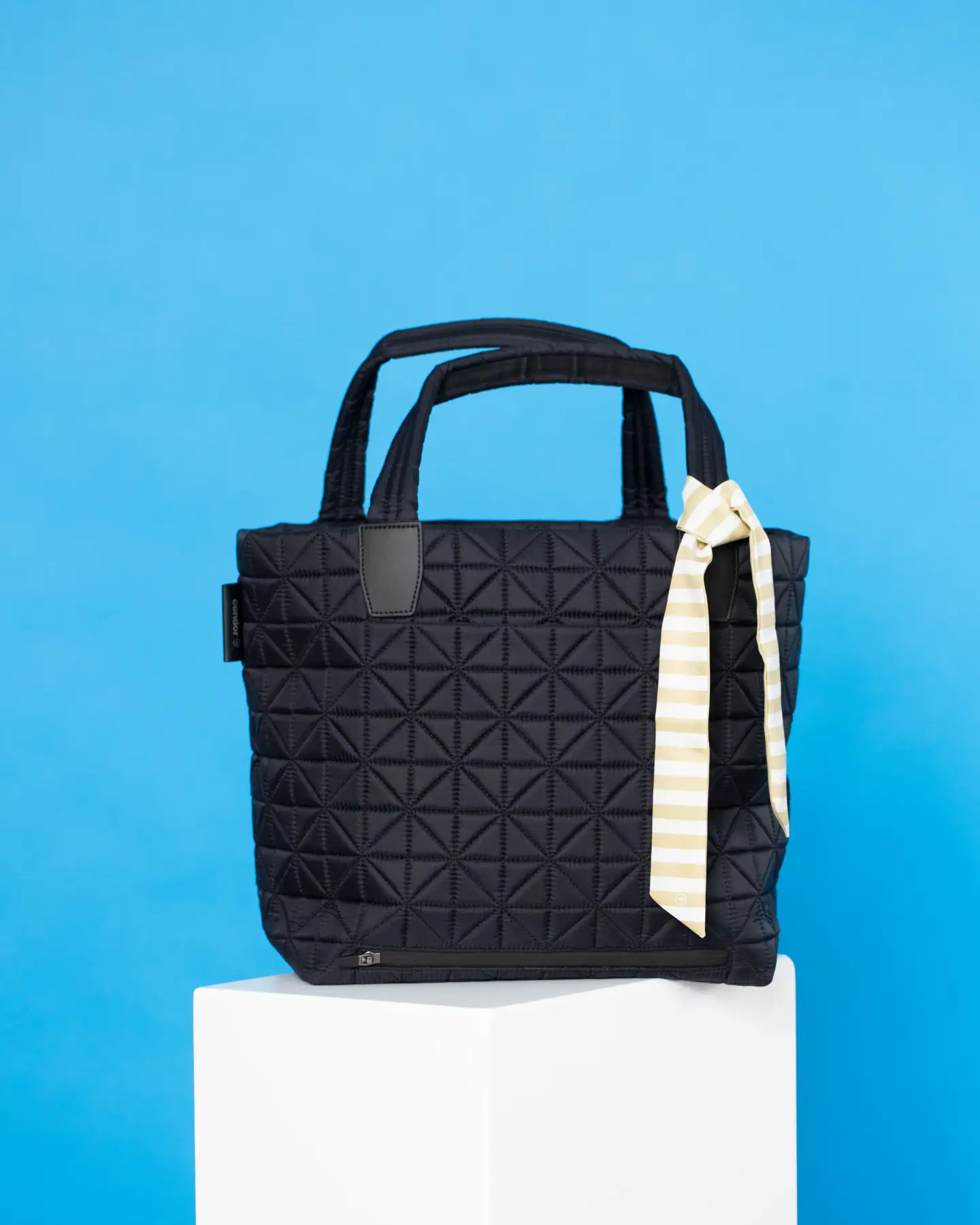 Una bolsa negra sobre un pedestal blanco contra un fondo azul, con un paño rayado que cuelga del asa