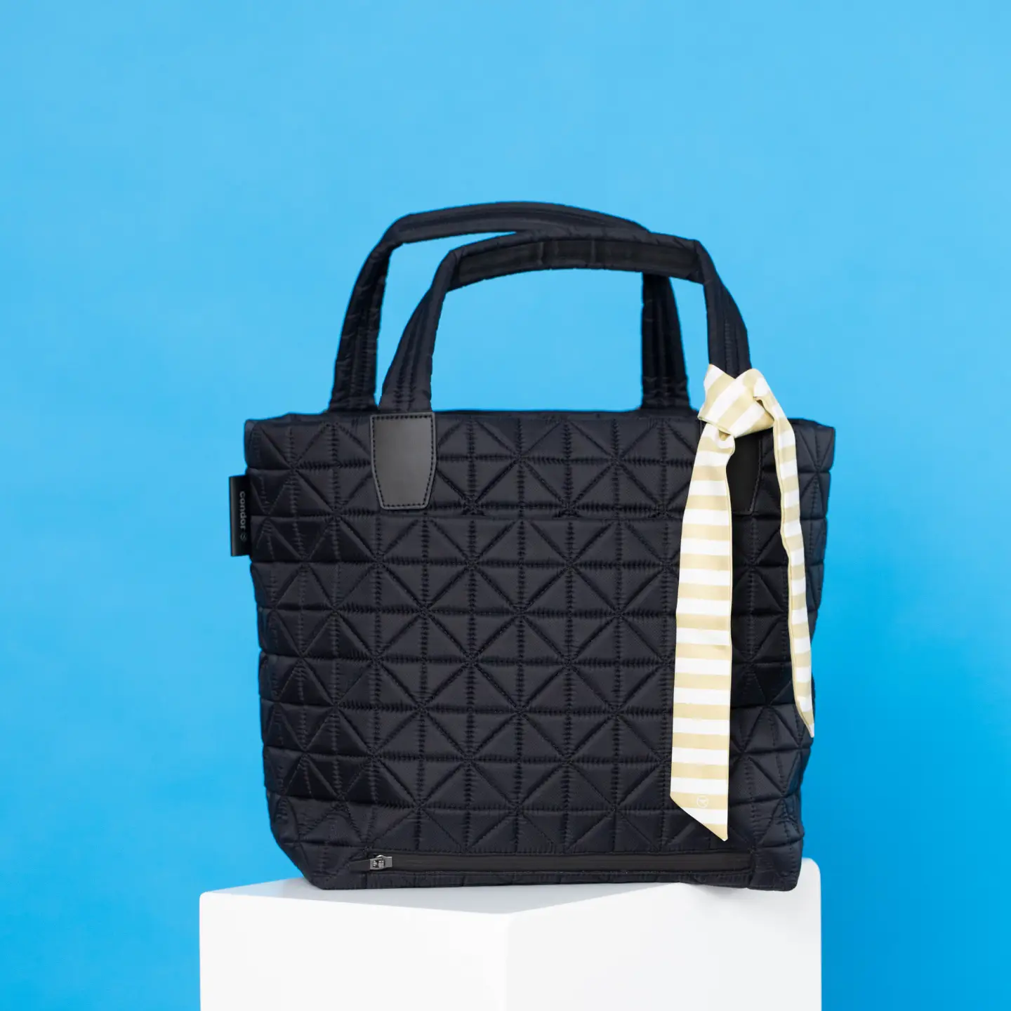 Una bolsa negra sobre un pedestal blanco contra un fondo azul, con un paño rayado que cuelga del asa