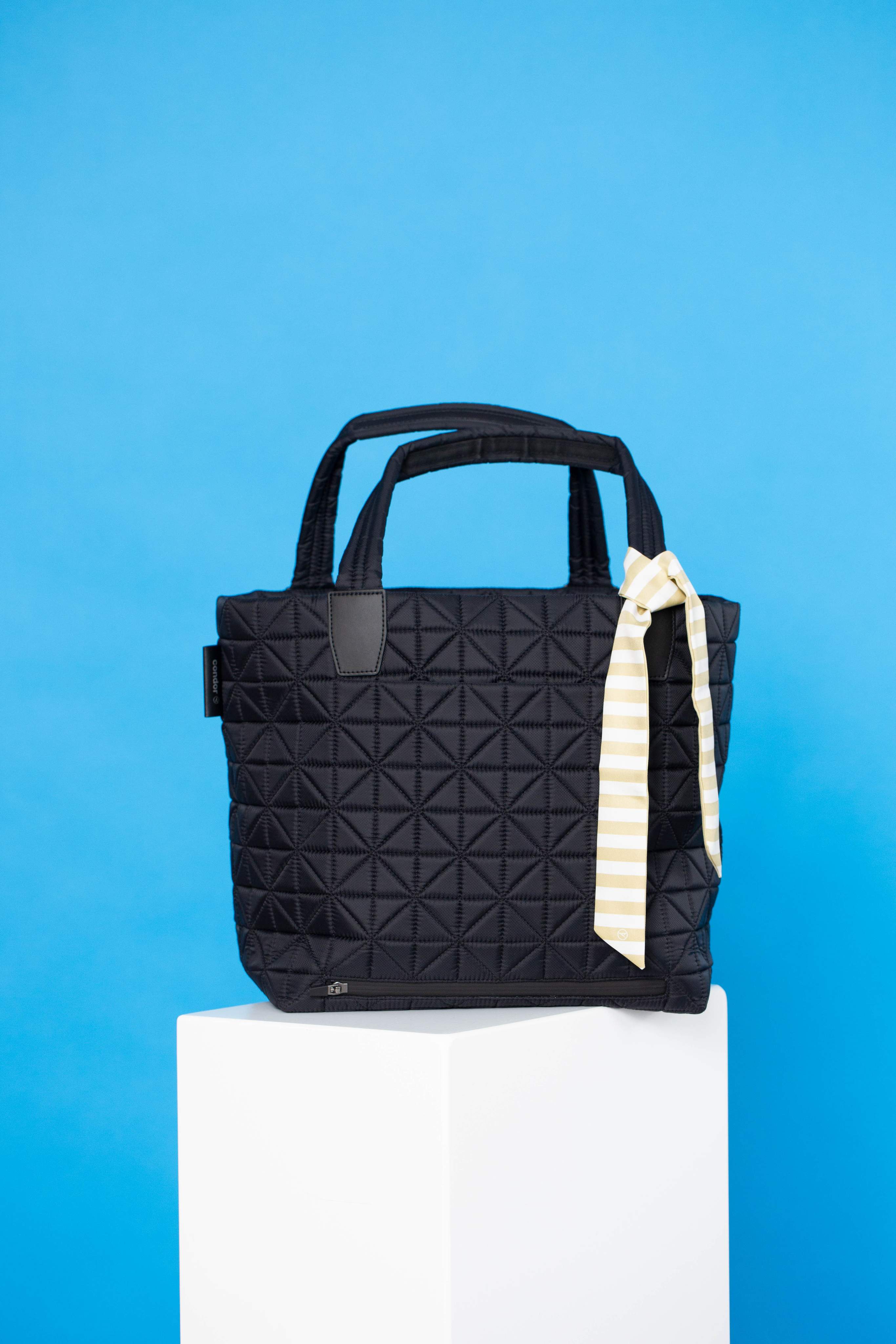 Un sac noir sur un piédestal blanc sur fond bleu, avec un tissu rayé suspendu à la poignée