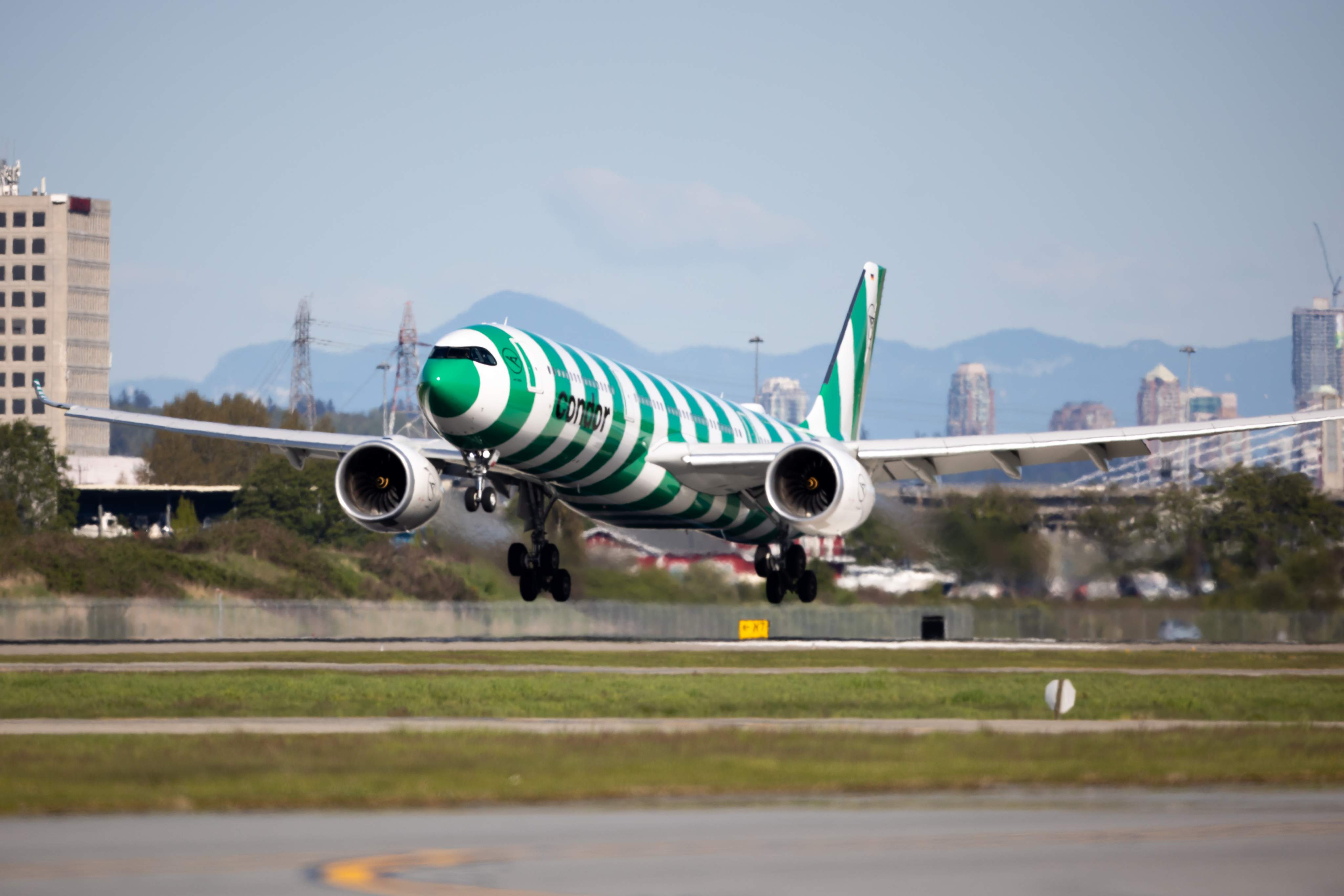 A green-striped Condor A330neo takes off from Vancouver Airport; the city skyline can be seen behind it, with mountains in the distance.