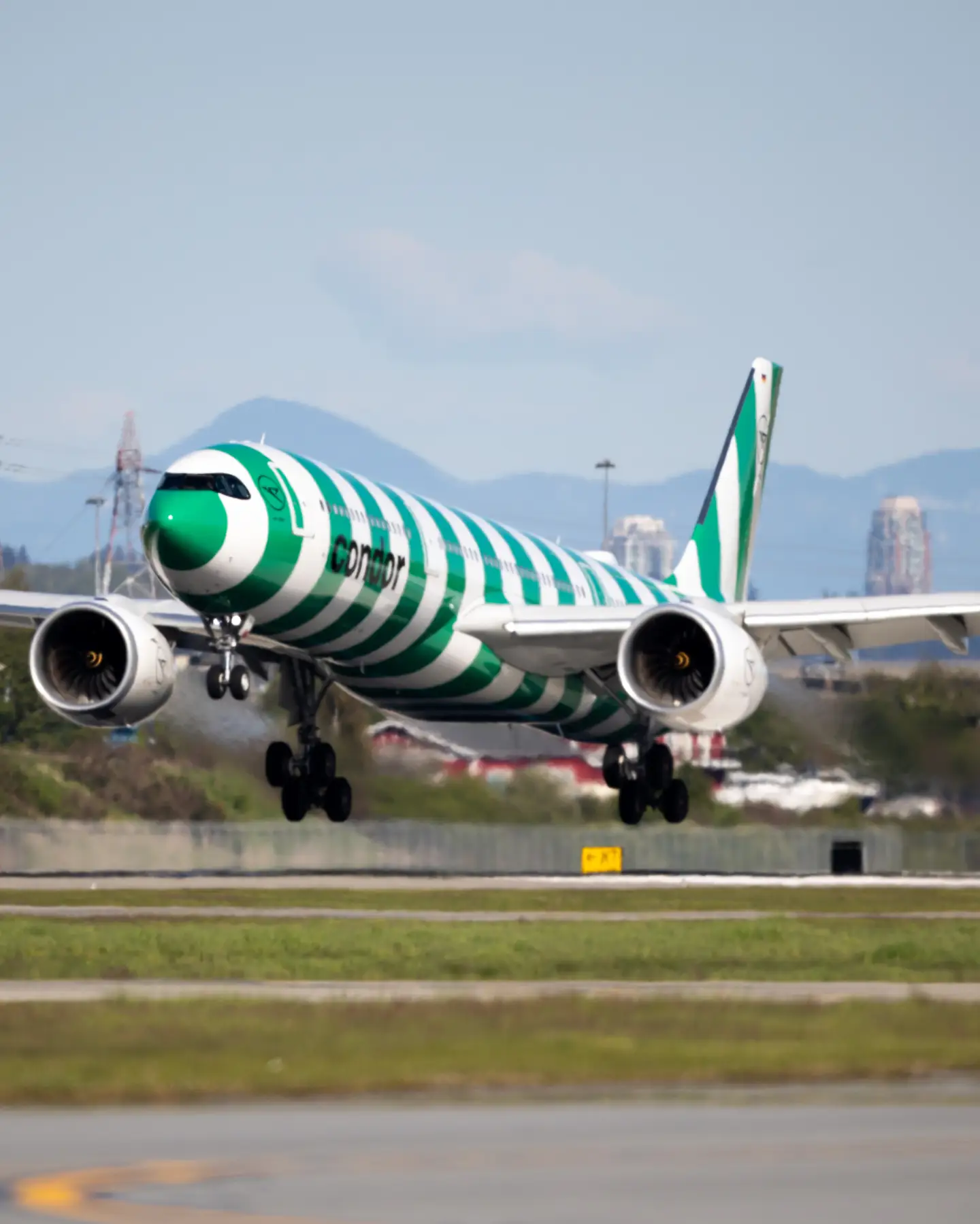 A green-striped Condor A330neo takes off from Vancouver Airport; the city skyline can be seen behind it, with mountains in the distance.