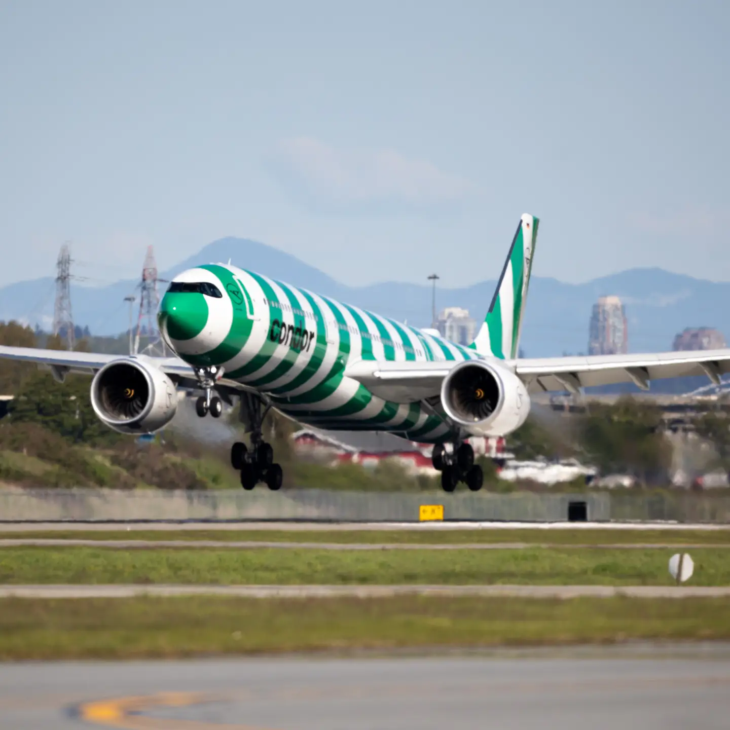 A green-striped Condor A330neo takes off from Vancouver Airport; the city skyline can be seen behind it, with mountains in the distance.