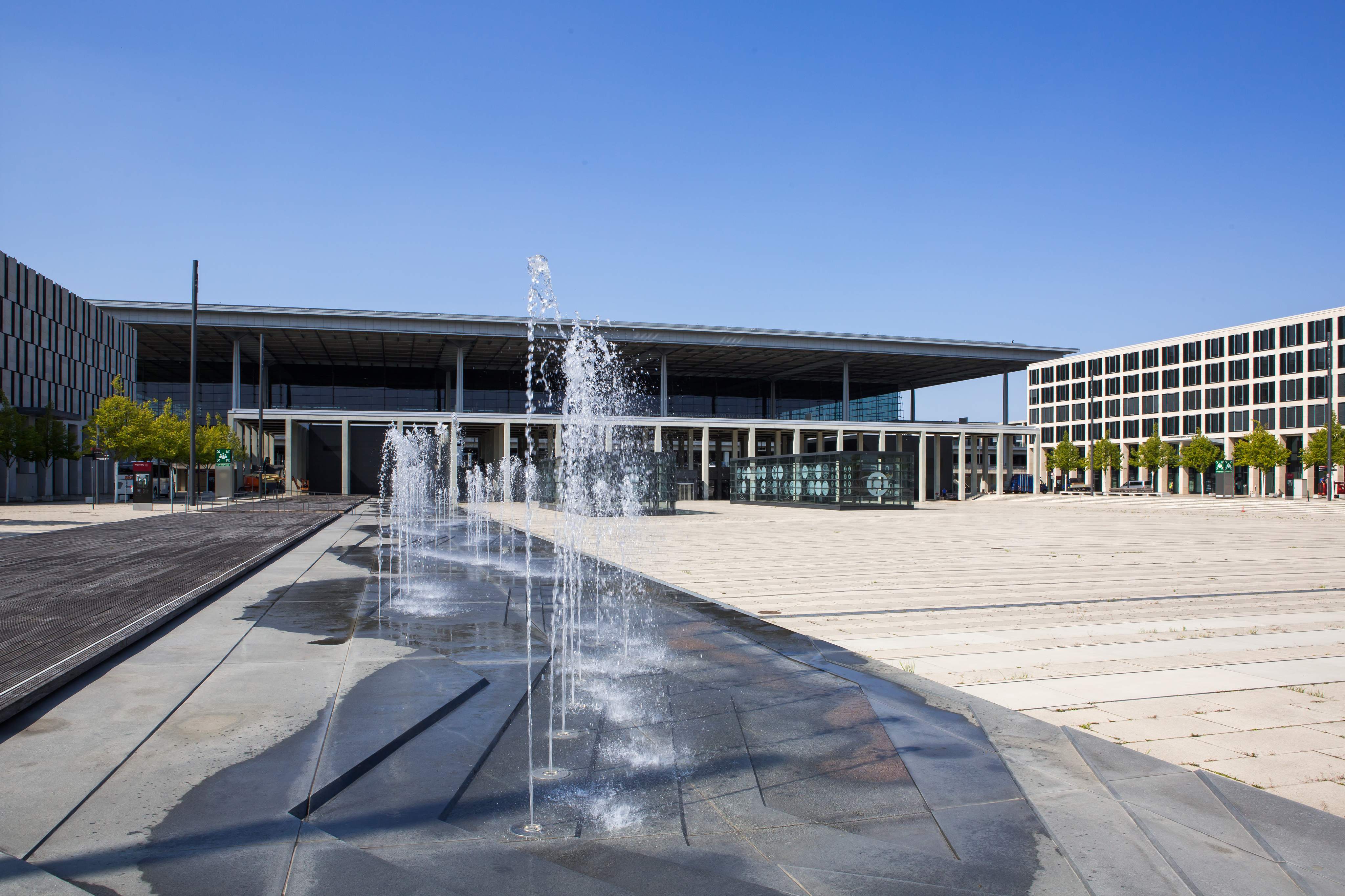 Vorplatz des Flughafens Berlin Brandenburg (BER) mit Wasserfontänen vor Terminal 1 bei klarem Himmel.