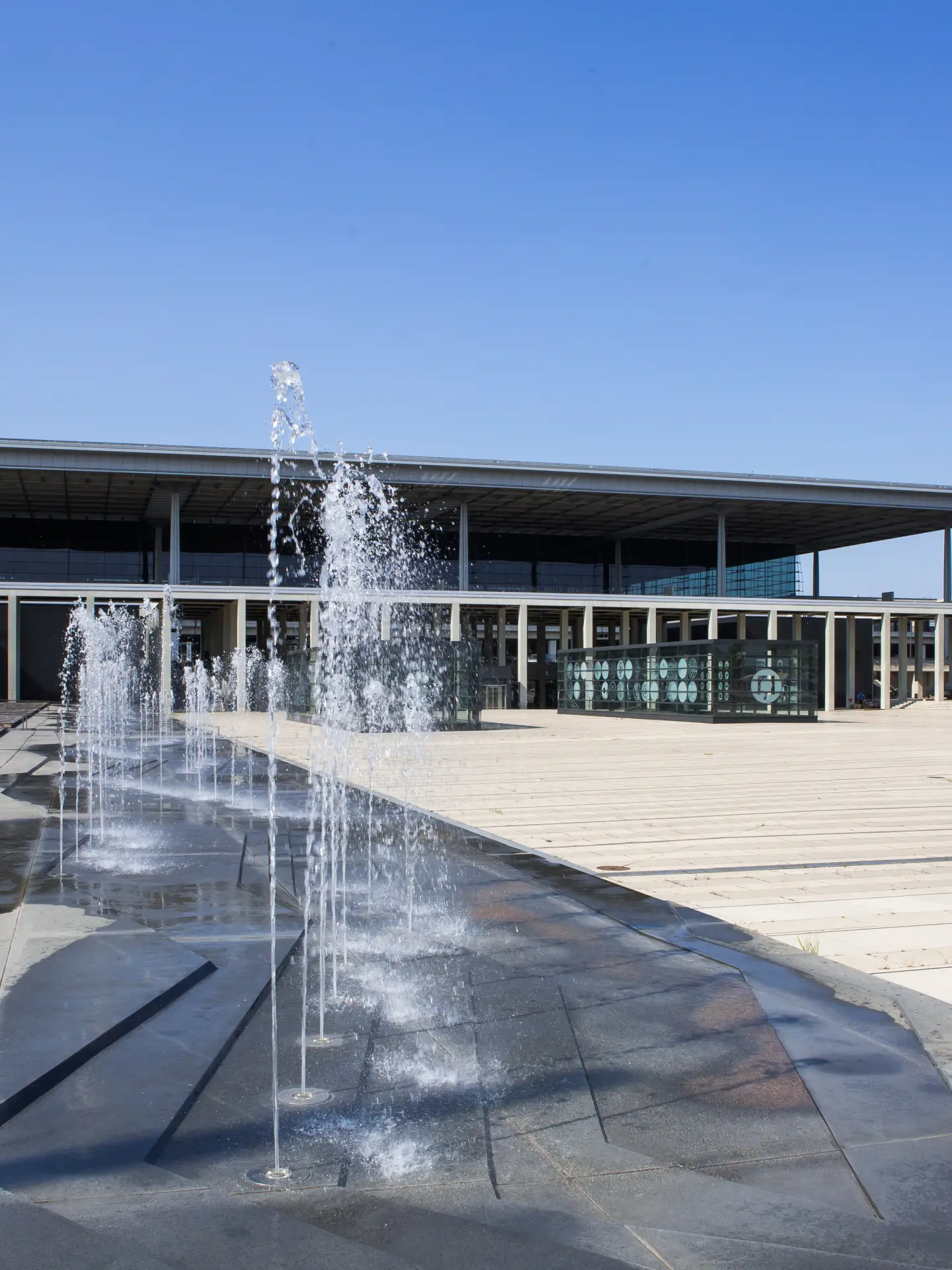Vorplatz des Flughafens Berlin Brandenburg (BER) mit Wasserfontänen vor Terminal 1 bei klarem Himmel.