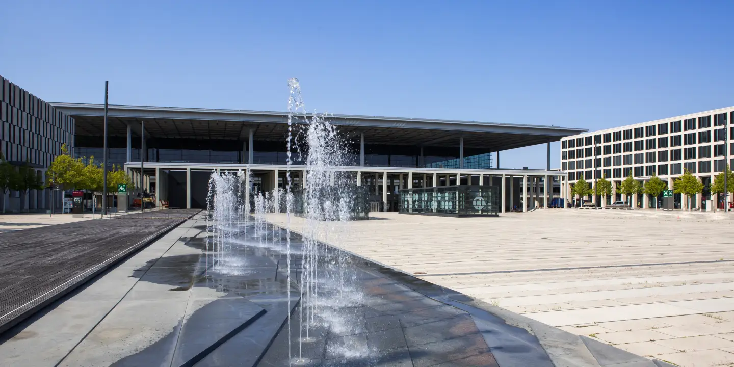 Vorplatz des Flughafens Berlin Brandenburg (BER) mit Wasserfontänen vor Terminal 1 bei klarem Himmel.