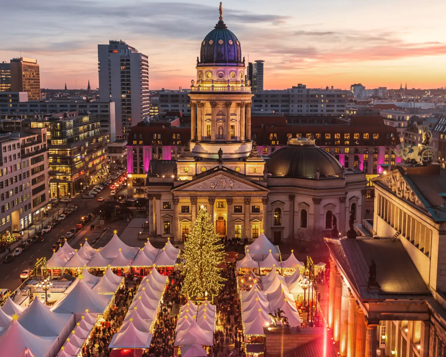 Festively lit Christmas market at Berlin’s Gendarmenmarkt, featuring a large Christmas tree and the French Cathedral at sunset.