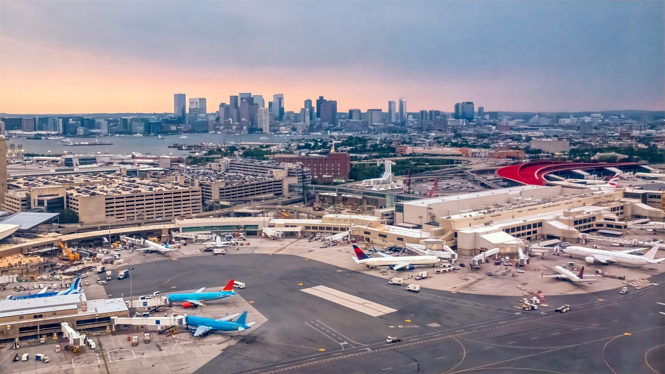 Aerial view of Boston Logan International Airport with terminals, aircraft, and the Boston skyline in the background.
