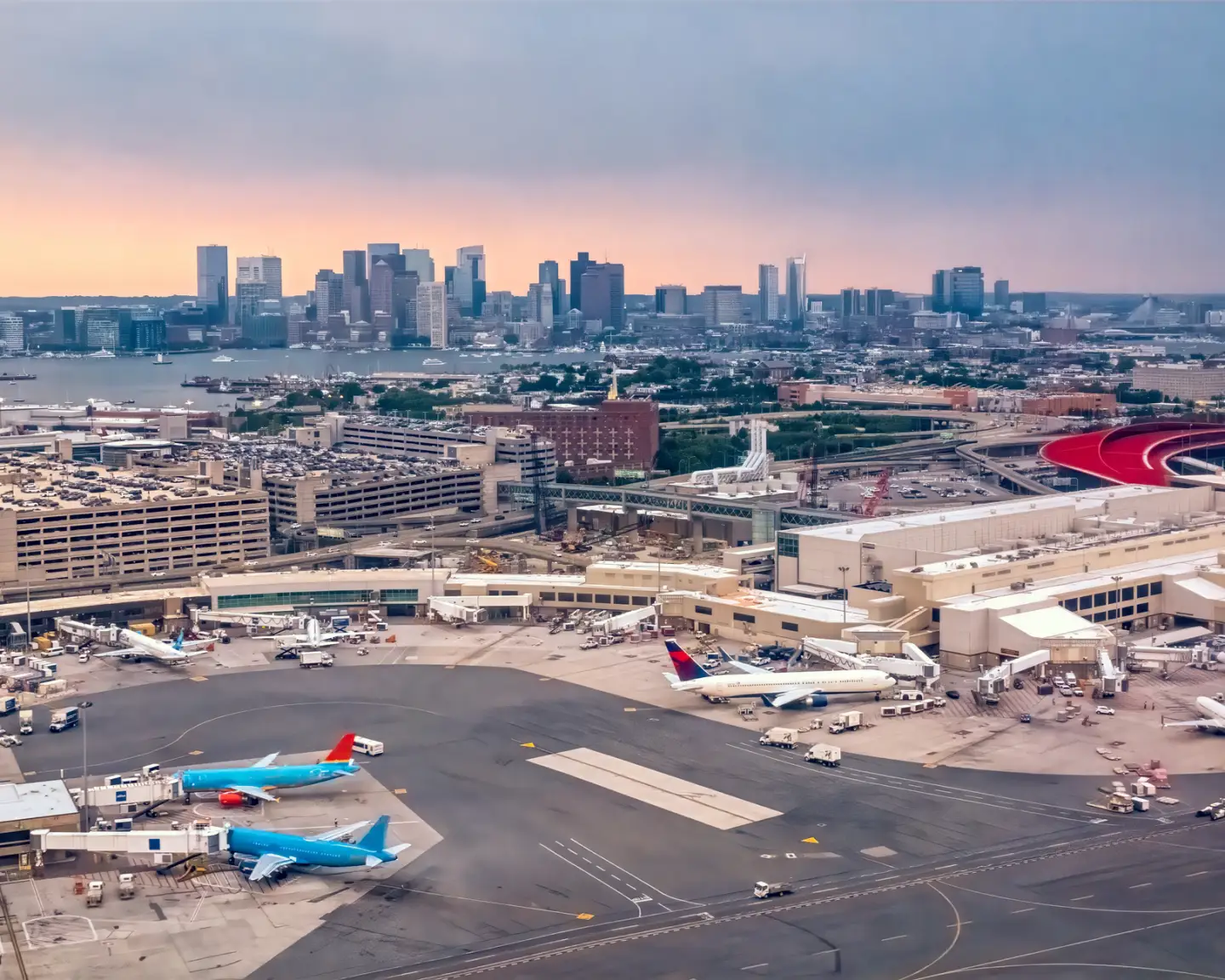 Aerial view of Boston Logan International Airport with terminals, aircraft, and the Boston skyline in the background.