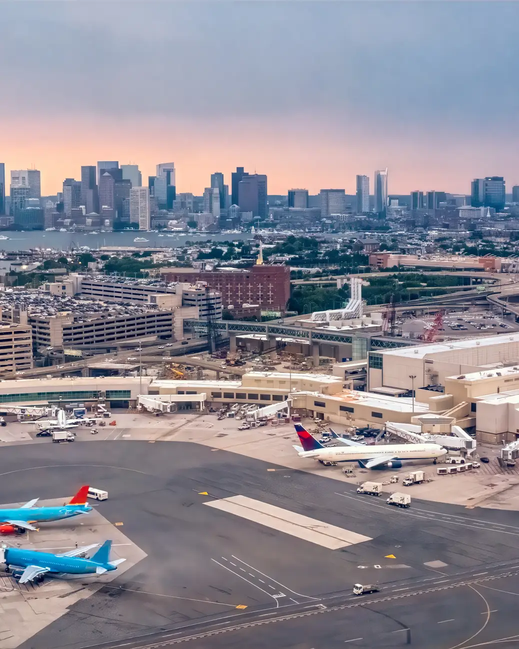 Aerial view of Boston Logan International Airport with terminals, aircraft, and the Boston skyline in the background.