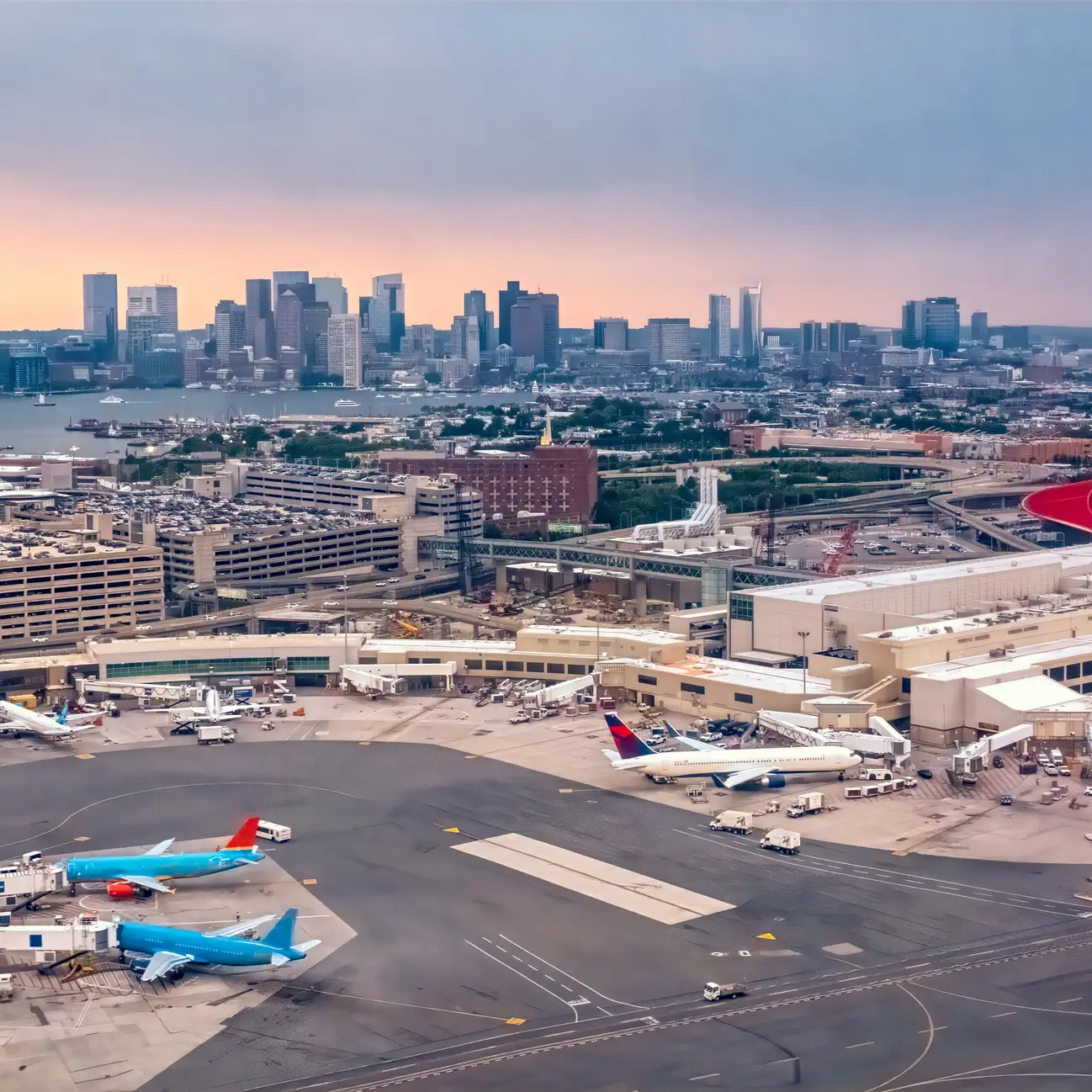 Aerial view of Boston Logan International Airport with terminals, aircraft, and the Boston skyline in the background.