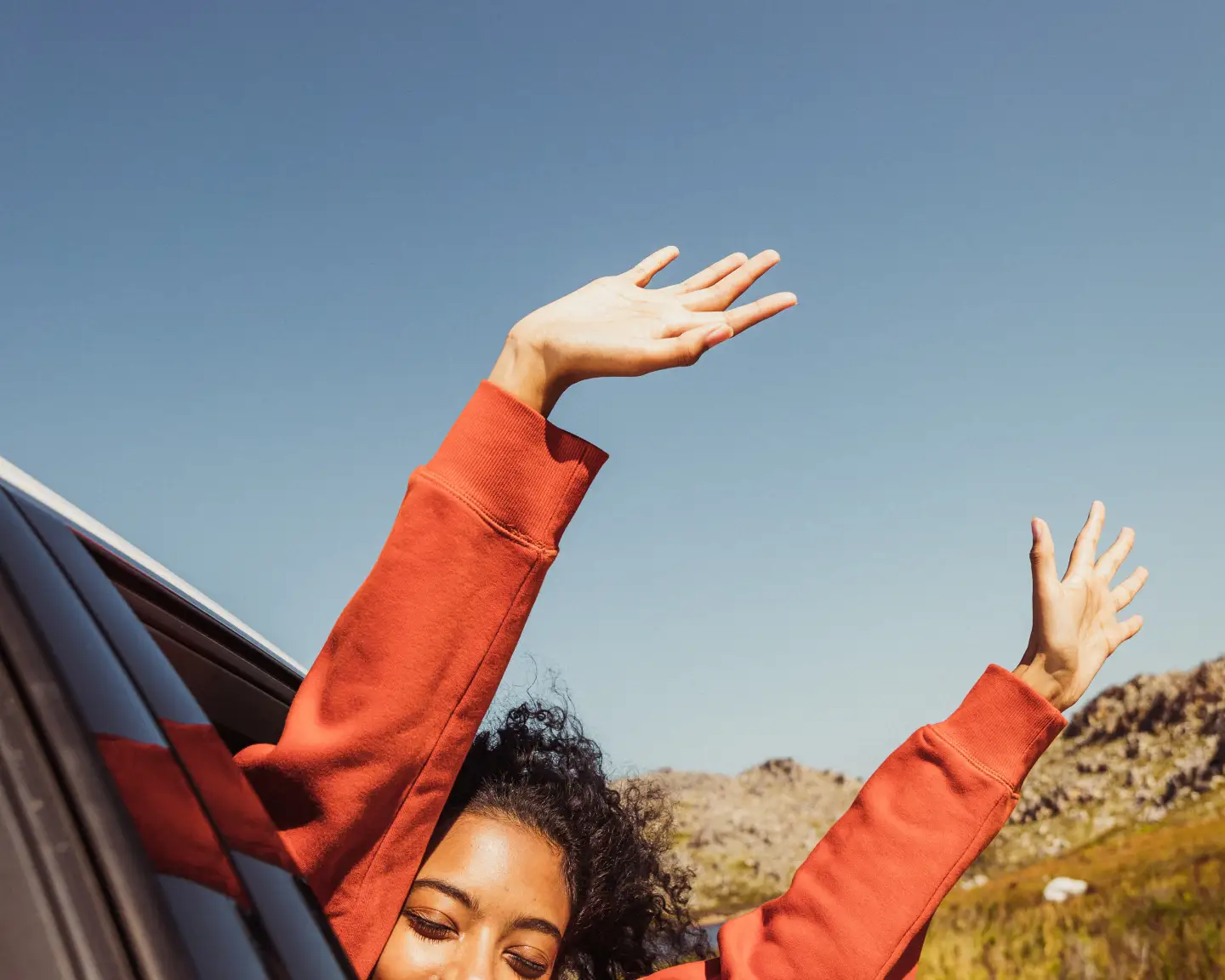 
A woman joyfully leaning out of a car window, with her arms raised in the air, enjoying a sunny day on a scenic road.
