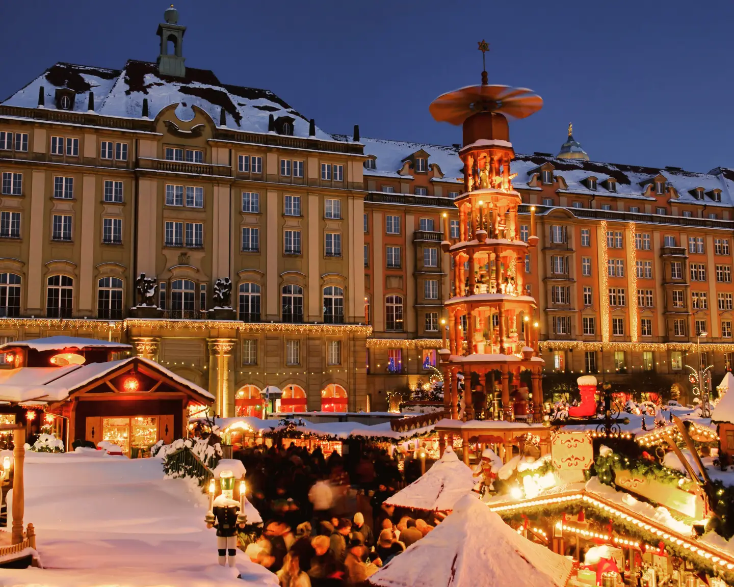 Snow-covered Dresden Striezelmarkt with festively lit Christmas market stalls and a large Christmas pyramid in the evening.