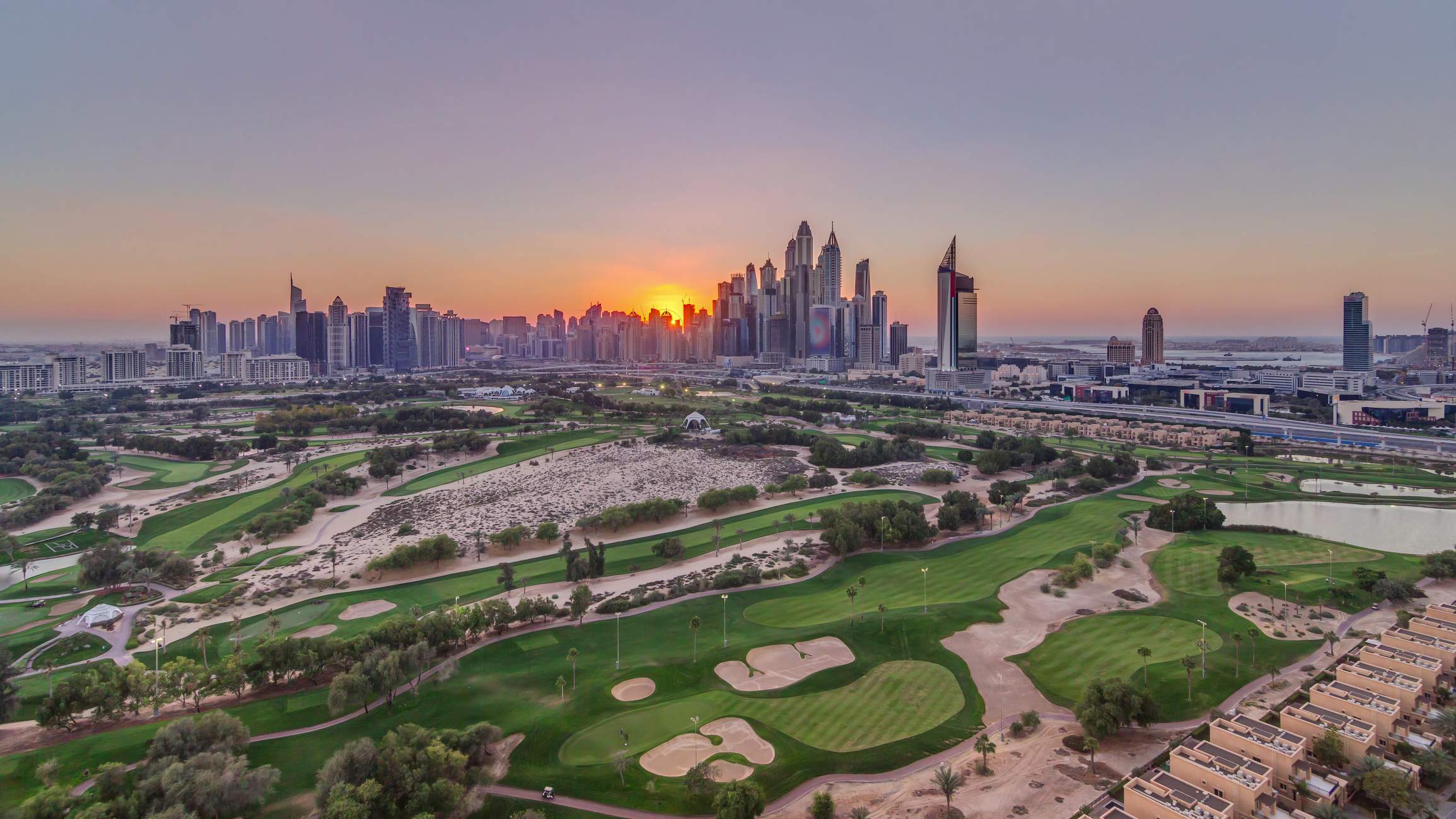 Blick über einen Golfplatz in Dubai bei Sonnenuntergang mit grünen Fairways und der Skyline im Hintergrund.