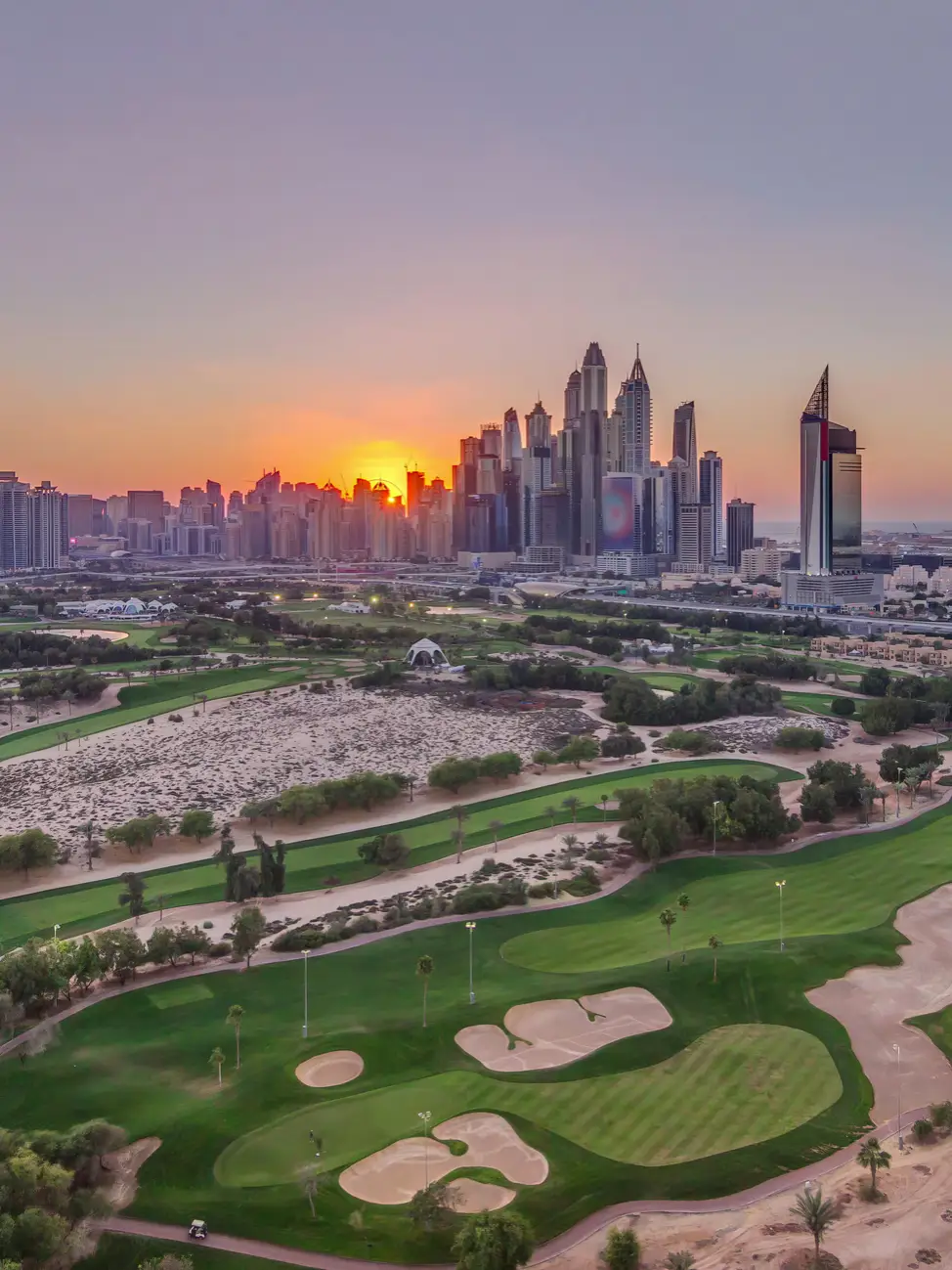 Blick über einen Golfplatz in Dubai bei Sonnenuntergang mit grünen Fairways und der Skyline im Hintergrund.