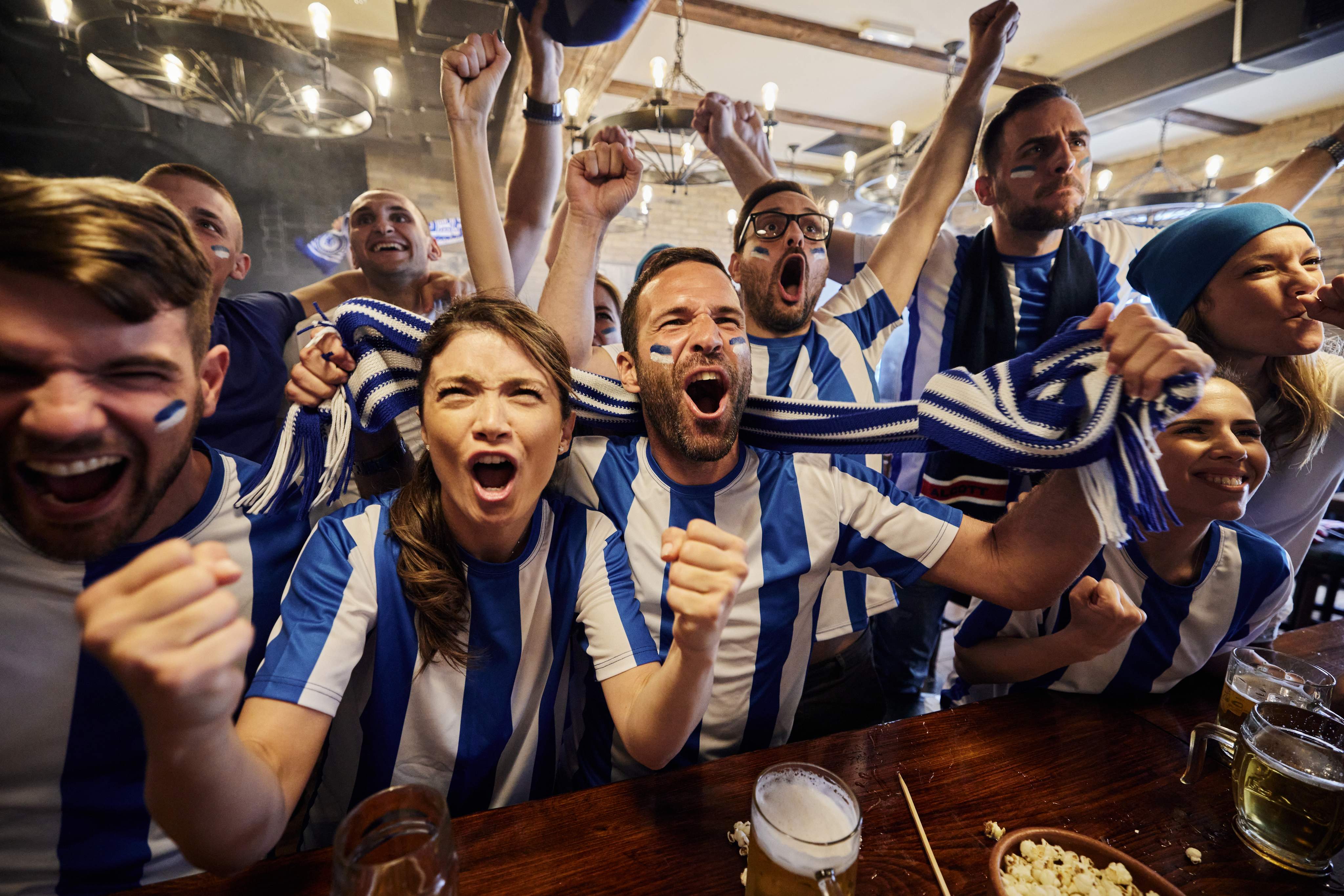 Large group of cheerful sports fans screaming while celebrating the success of their team during a game on TV in a bar.
