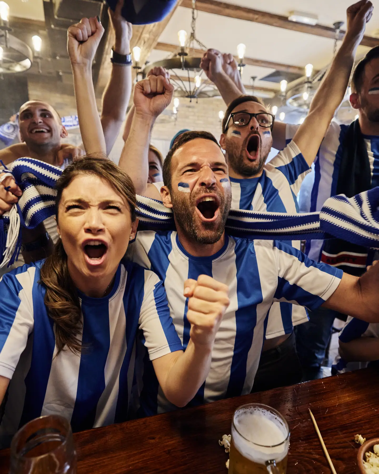 Large group of cheerful sports fans screaming while celebrating the success of their team during a game on TV in a bar.