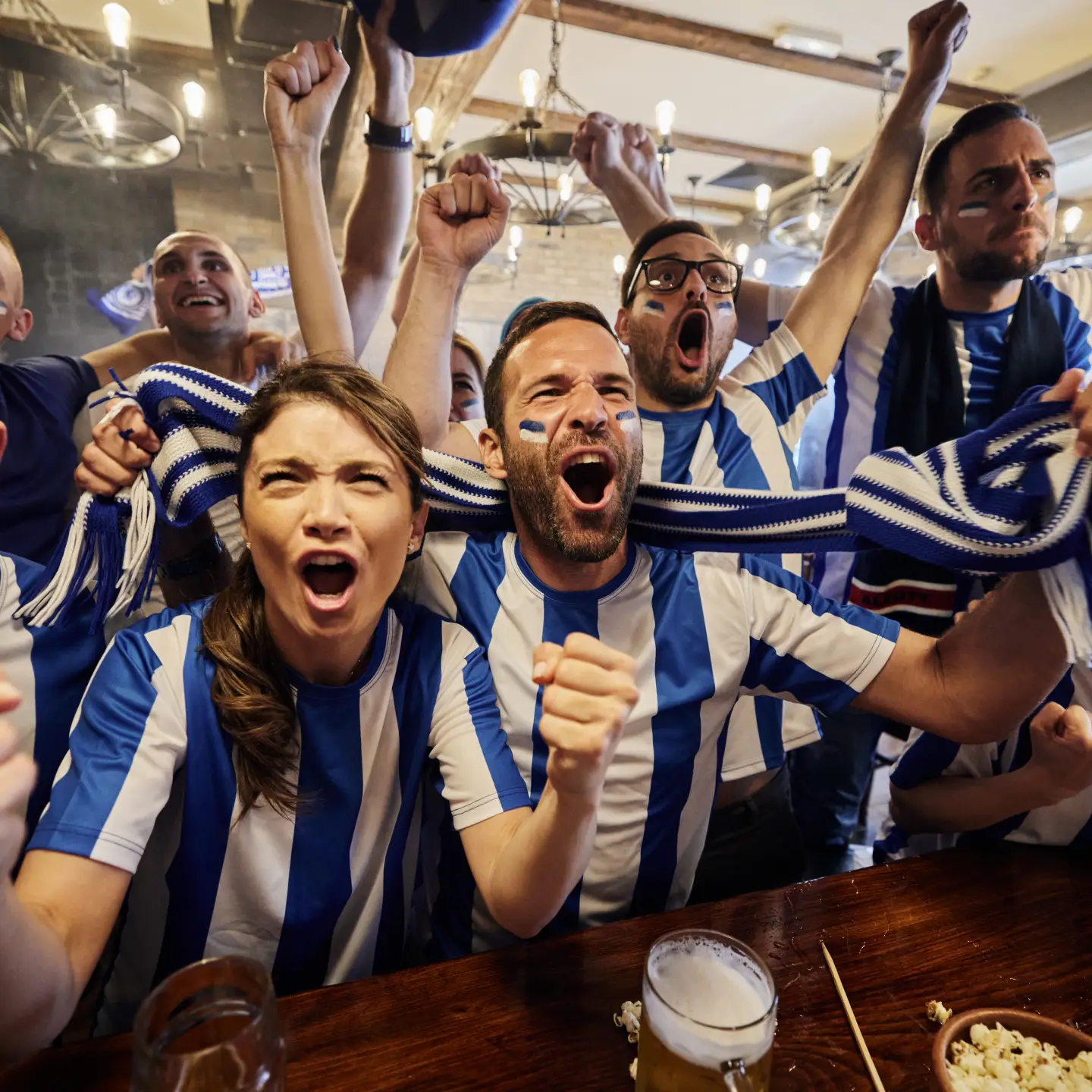Large group of cheerful sports fans screaming while celebrating the success of their team during a game on TV in a bar.