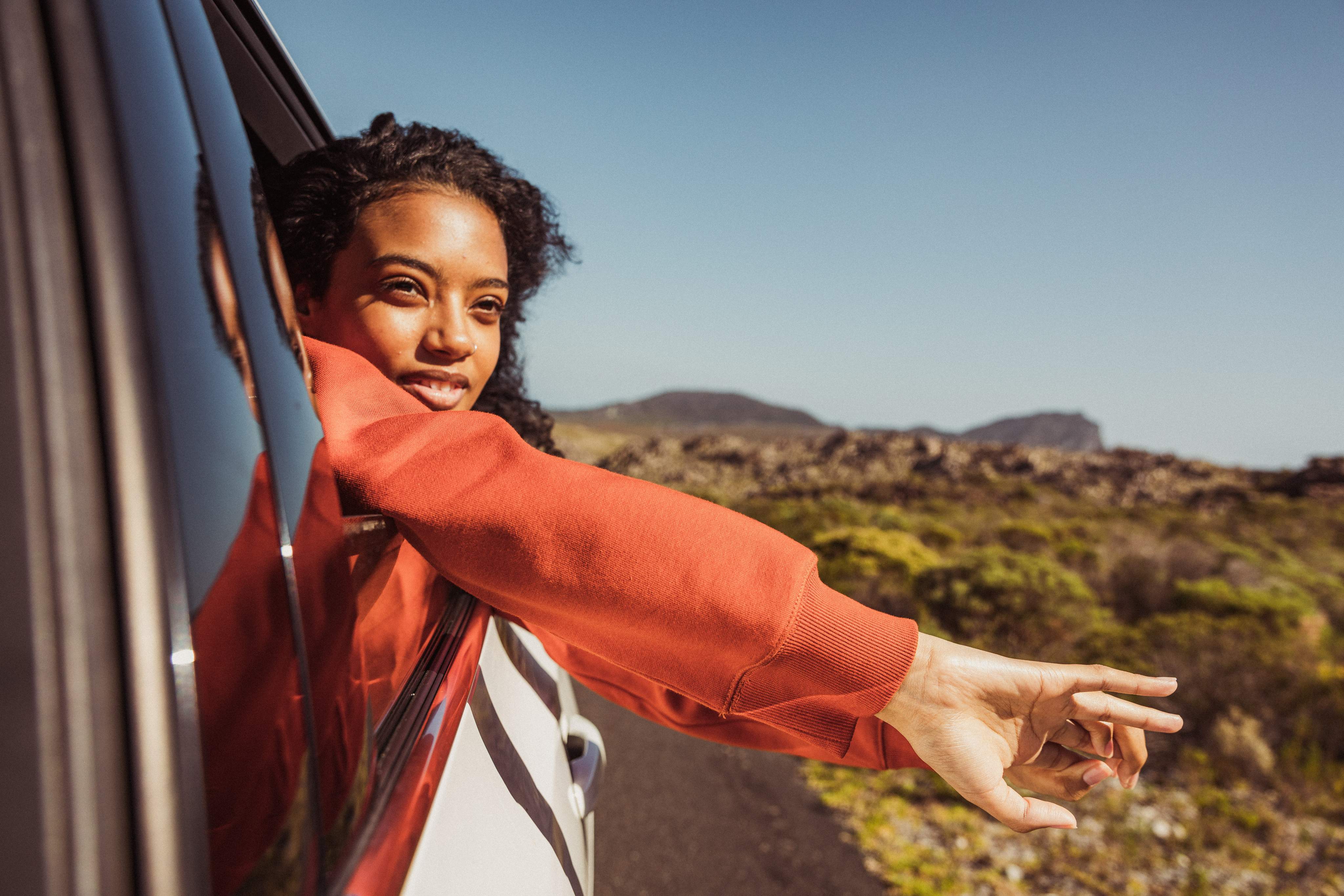 Une femme regarde par la fenêtre d'une voiture, ses bras pendent négligemment par-dessus la portière ; à l'arrière-plan, on aperçoit un paysage désertique.