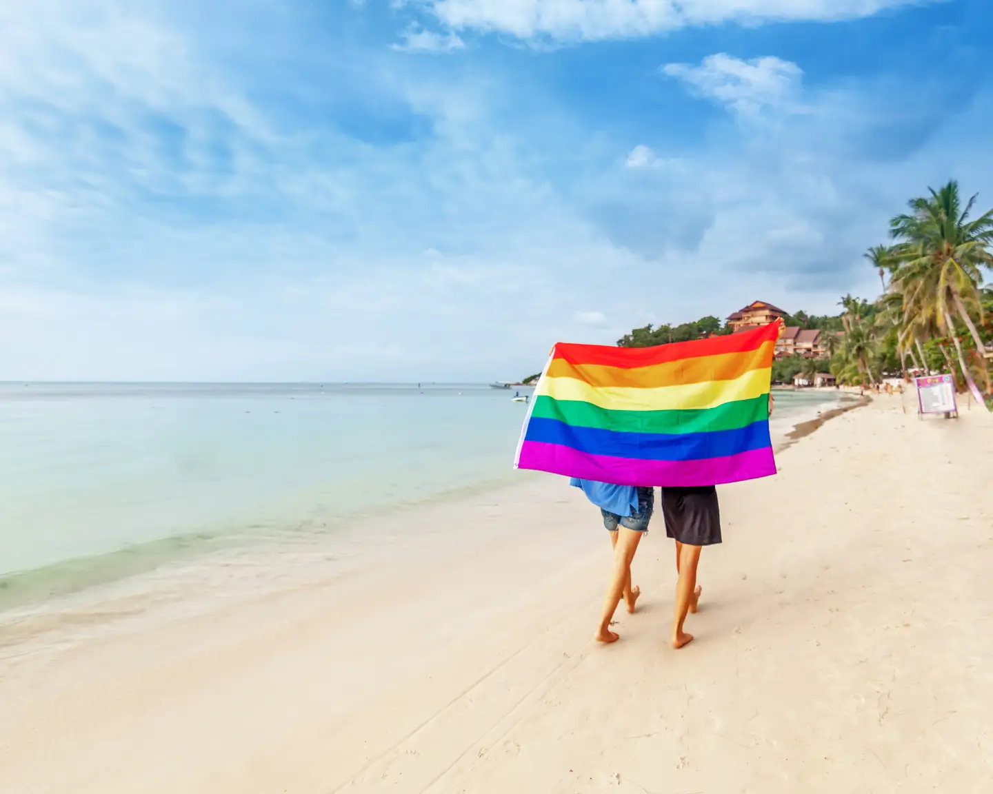 Ein junges lesbisches Paar spaziert an einem sonnigen Tropenstrand entlang, an dem eine Regenbogenflagge weht – Symbol für Gleichberechtigung und die LGBTQ+-Community.