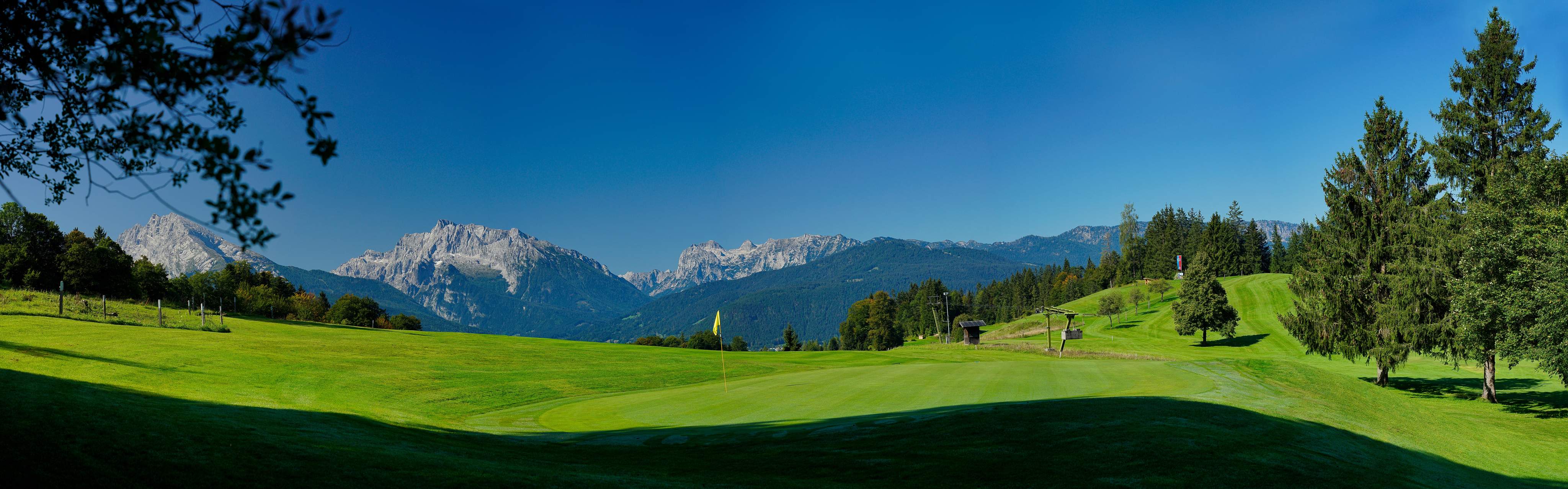 Panoramic view of a green golf course near Munich with a well-maintained fairway, surrounded by trees and the Alps in the background.