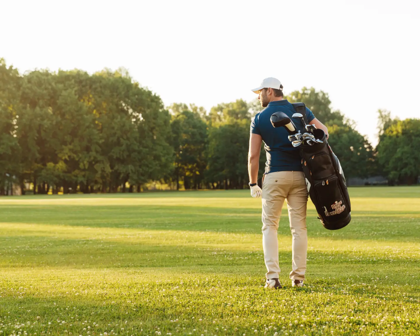Back view of young man in cap carrying golf bag while standing on field