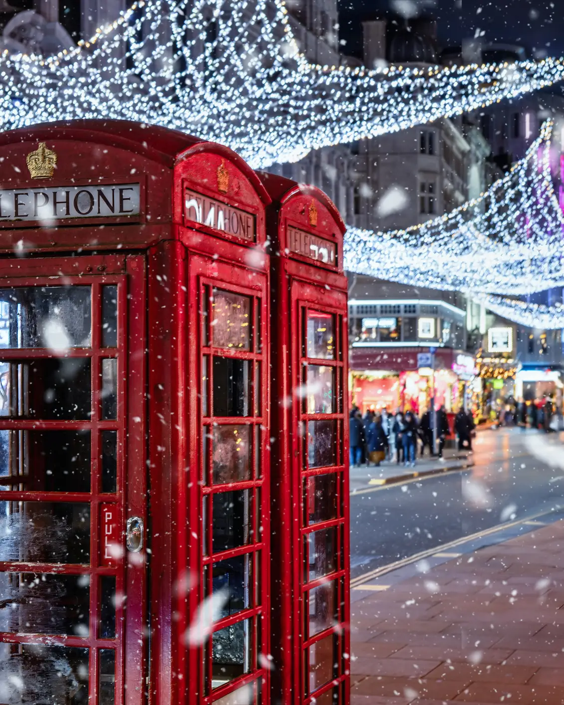 Rote Telefonzellen in London unter festlicher Weihnachtsbeleuchtung bei winterlichem Schneefall am Abend.