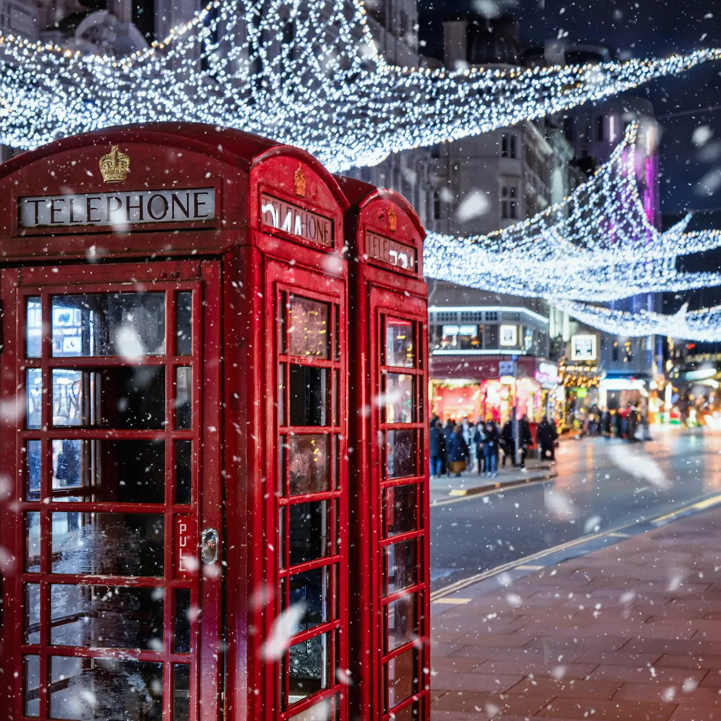 Rote Telefonzellen in London unter festlicher Weihnachtsbeleuchtung bei winterlichem Schneefall am Abend.