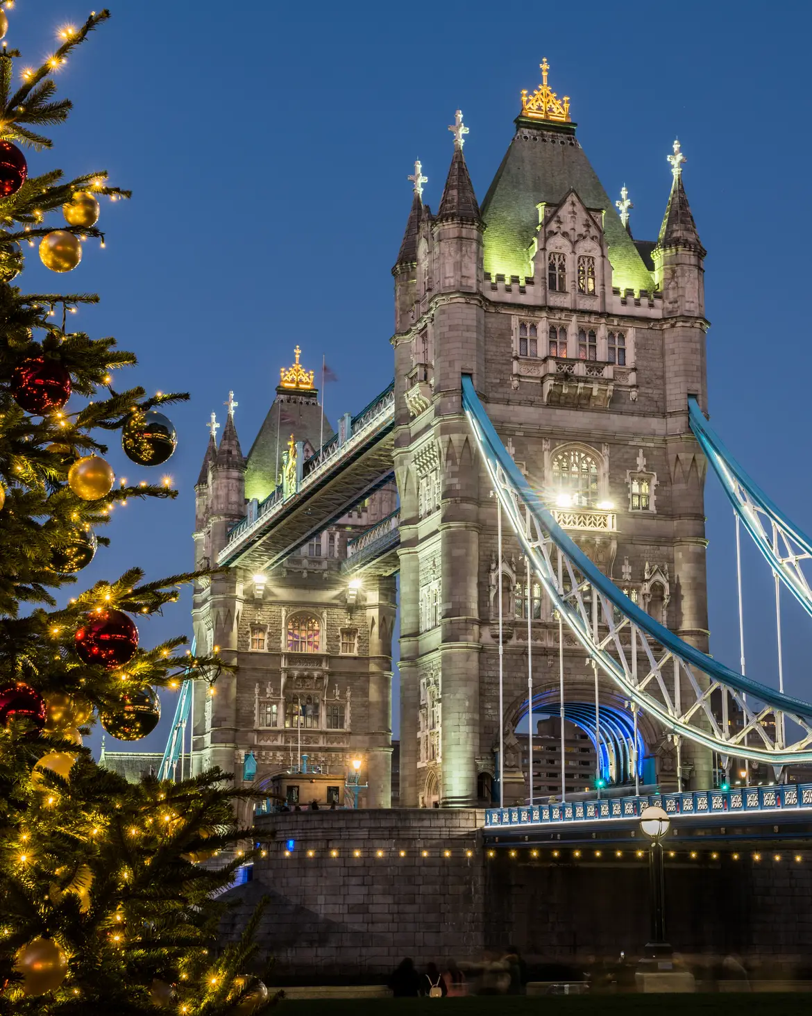 Beleuchtete Tower Bridge in London neben einem festlich geschmückten Weihnachtsbaum am Abend.