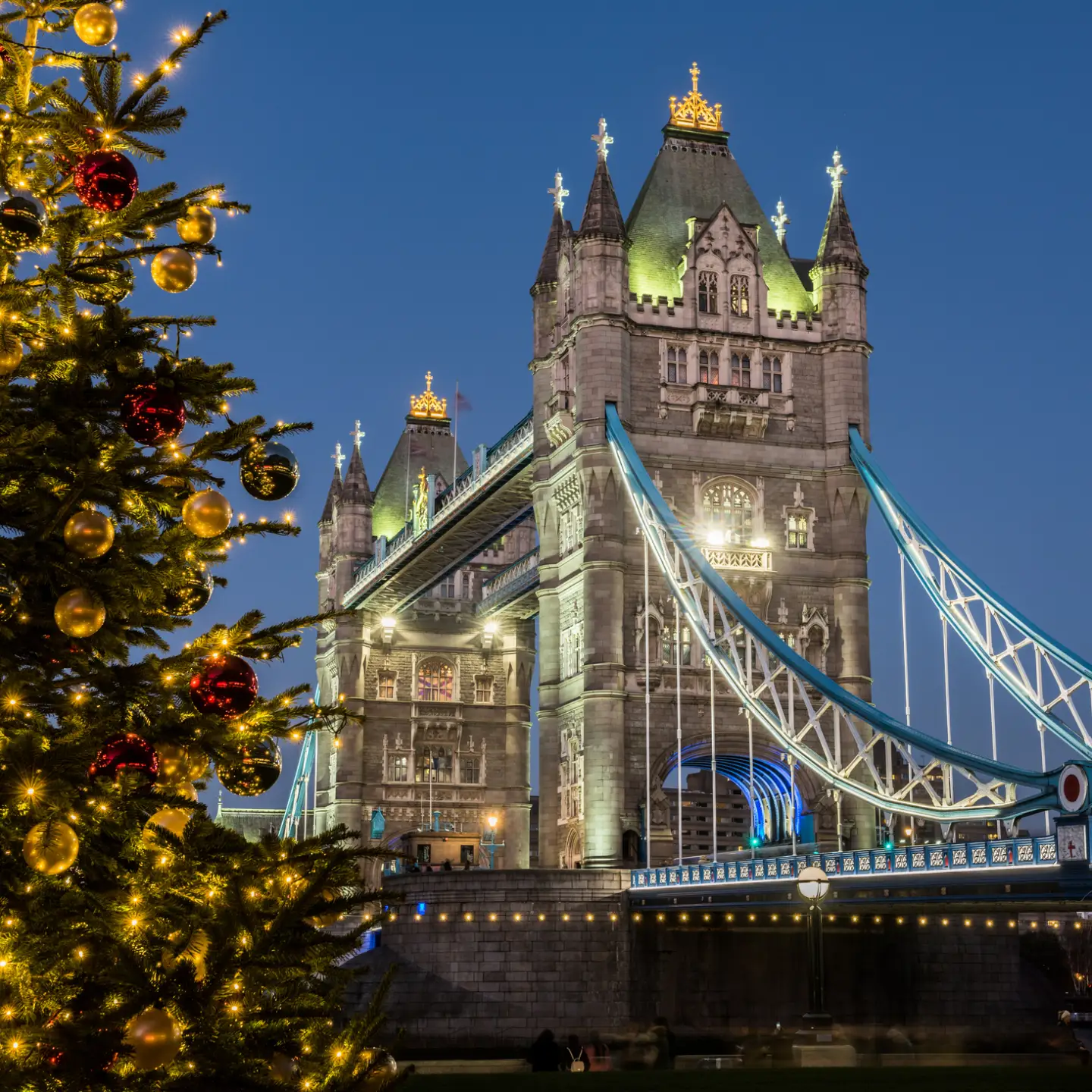 Beleuchtete Tower Bridge in London neben einem festlich geschmückten Weihnachtsbaum am Abend.