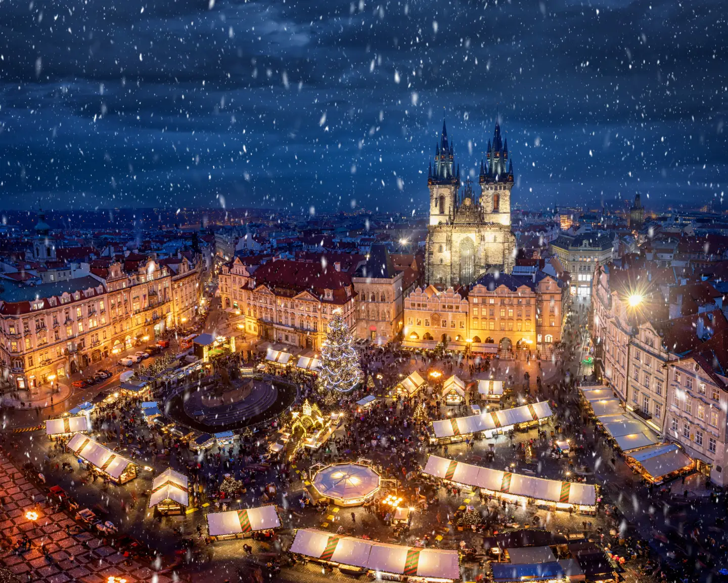 Festively lit Christmas market in the Old Town Square in Prague with a Christmas tree and the Church of Our Lady before Týn in snowfall.