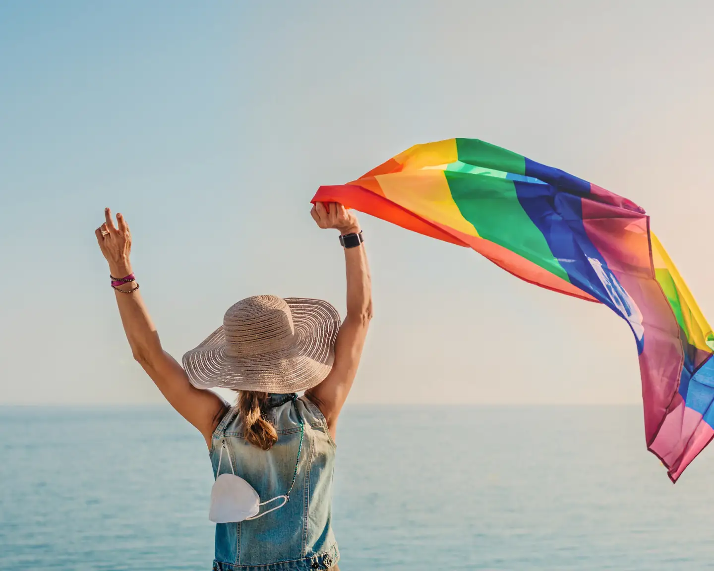Frau mit Sonnenhut hält am Strand eine Regenbogenflagge in die Höhe.
