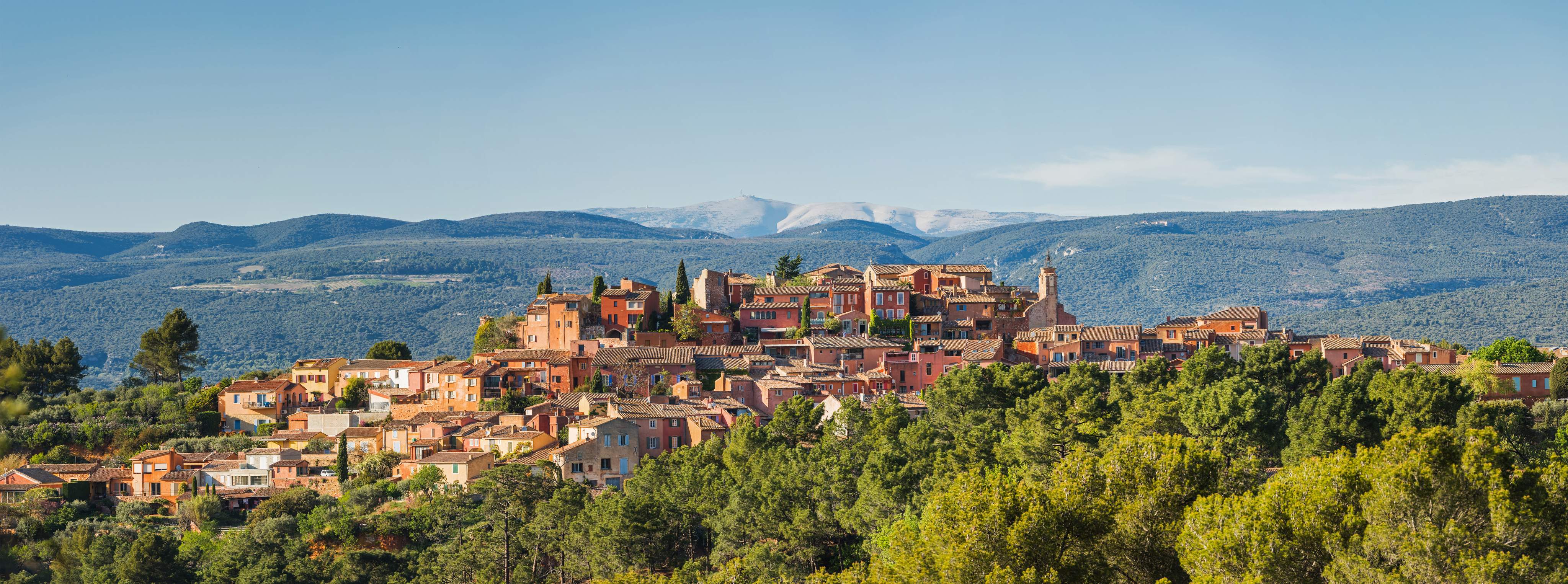 Panoramic view of the Provençal village of Roussillon with ocher-colored houses, green forests and the mountains of Provence in the background.