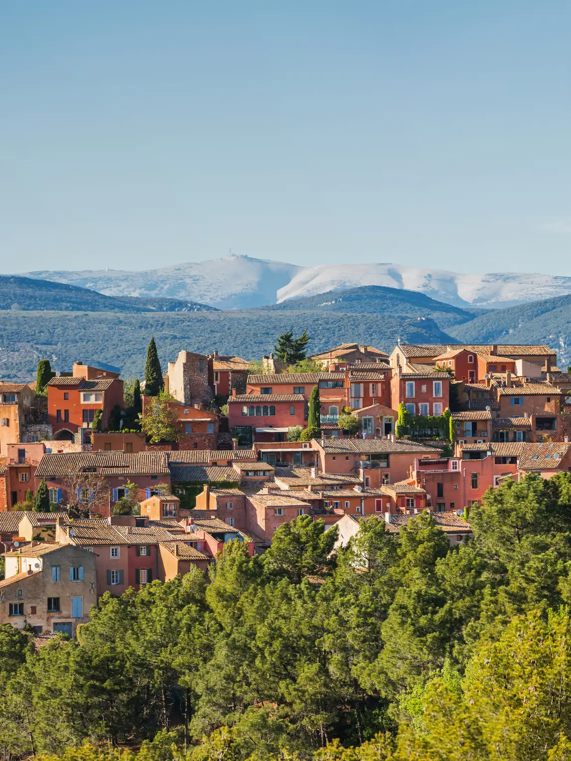 Panoramic view of the Provençal village of Roussillon with ocher-colored houses, green forests and the mountains of Provence in the background.