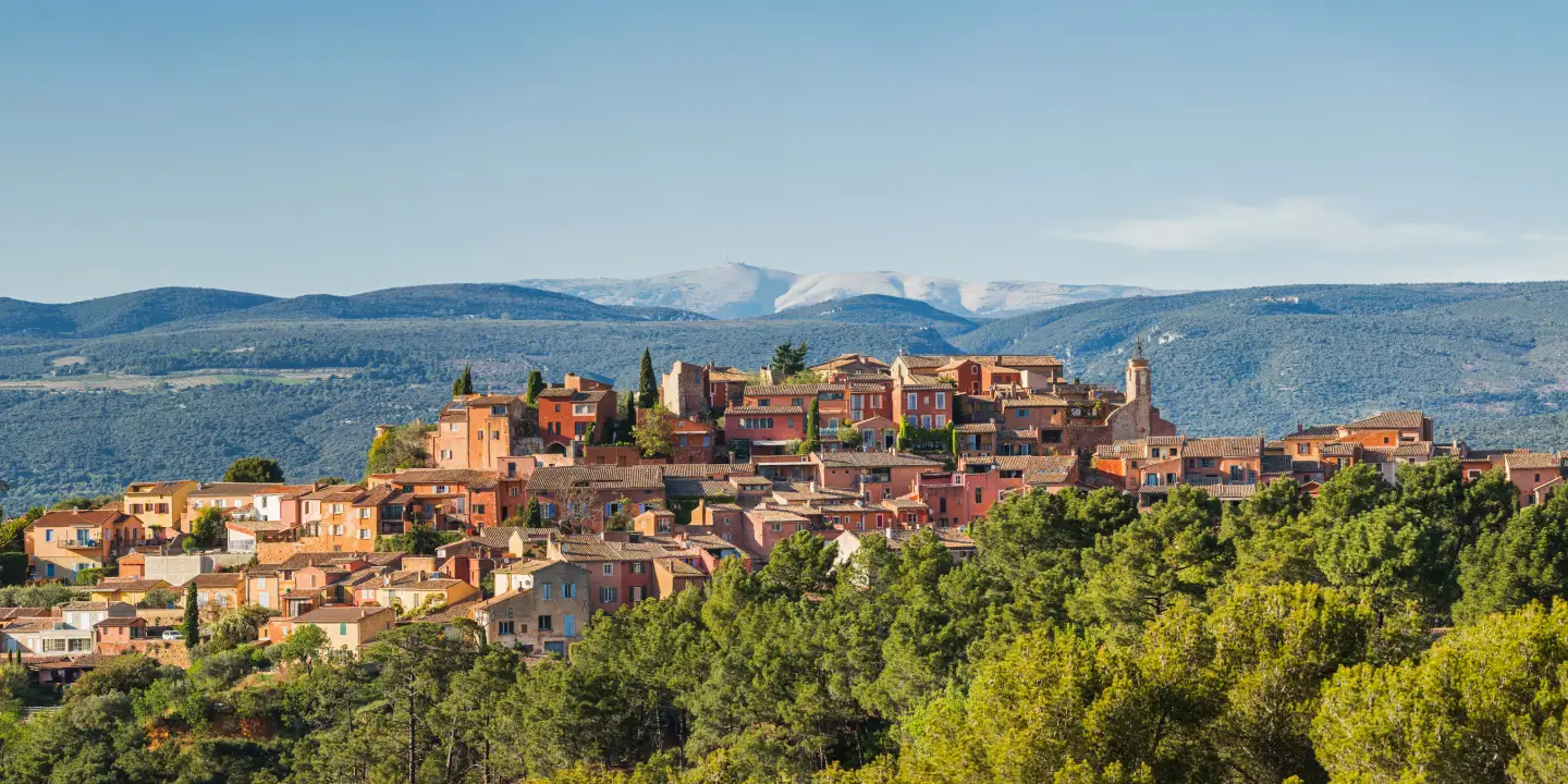 Panoramic view of the Provençal village of Roussillon with ocher-colored houses, green forests and the mountains of Provence in the background.