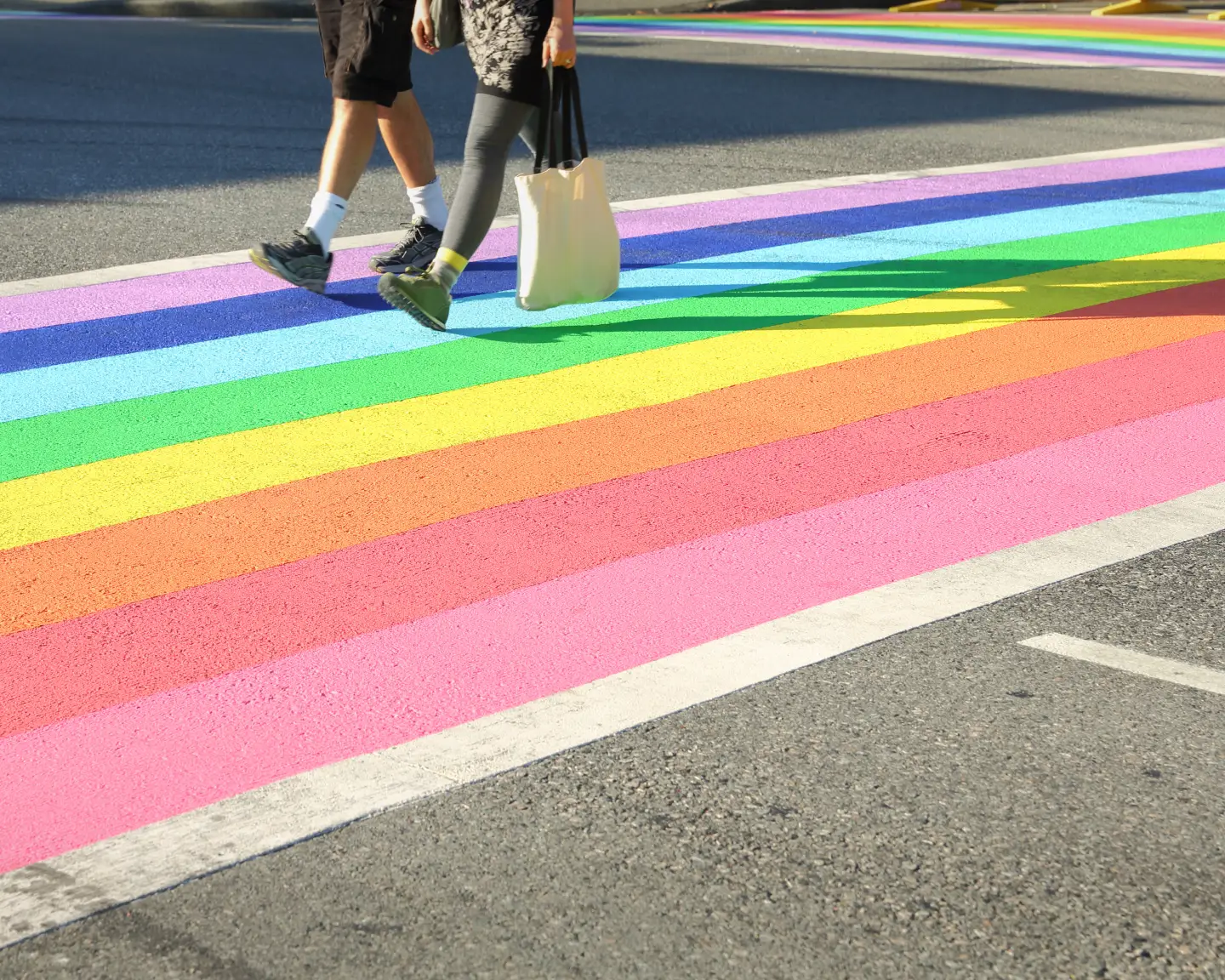Zwei Personen überqueren eine Straße auf einem leuchtend regenbogenfarbenen Zebrastreifen.