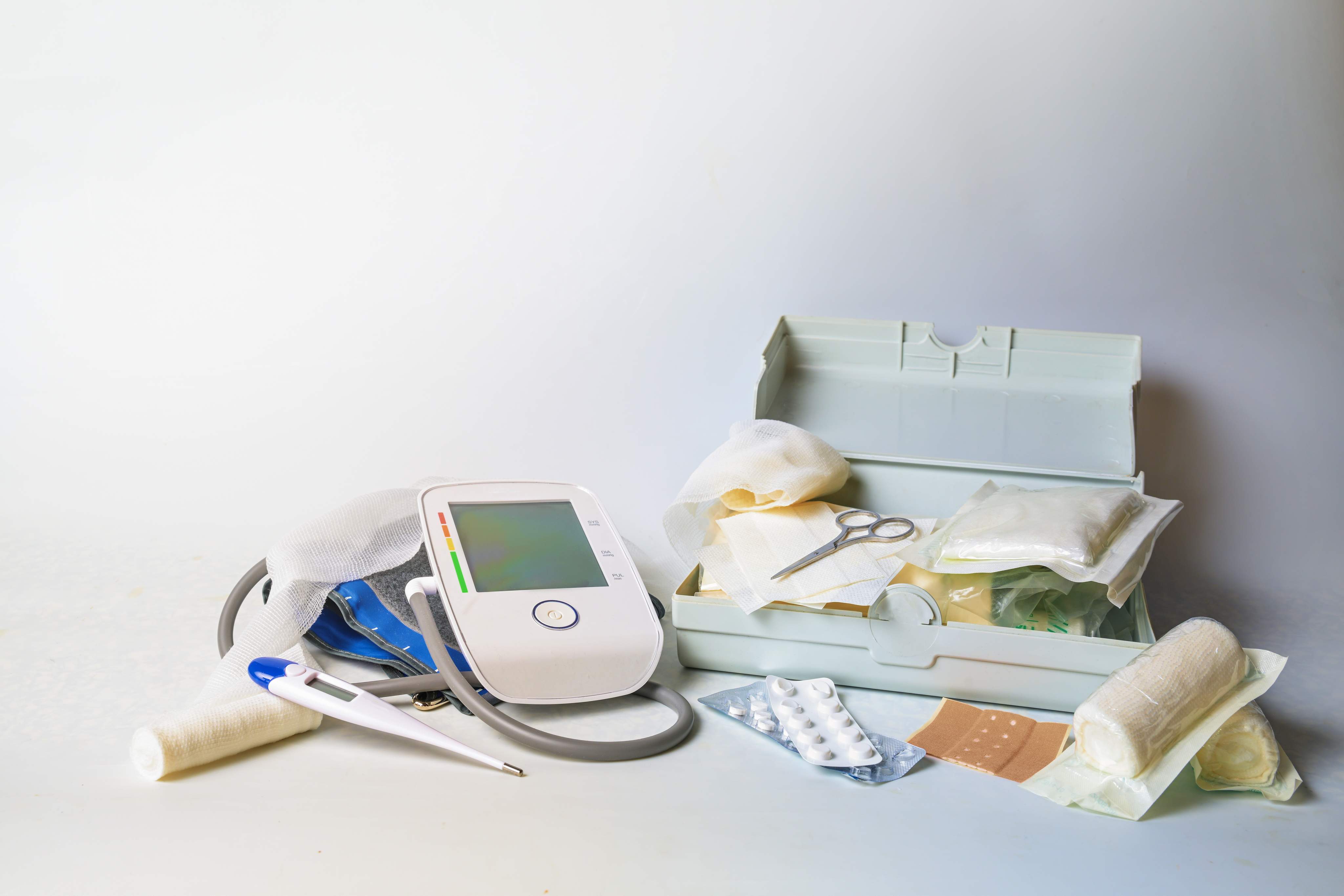 Blutdruckmessgerät, Thermometer, Medikamente und Verbandsmaterial liegen auf einer hellen Fläche.