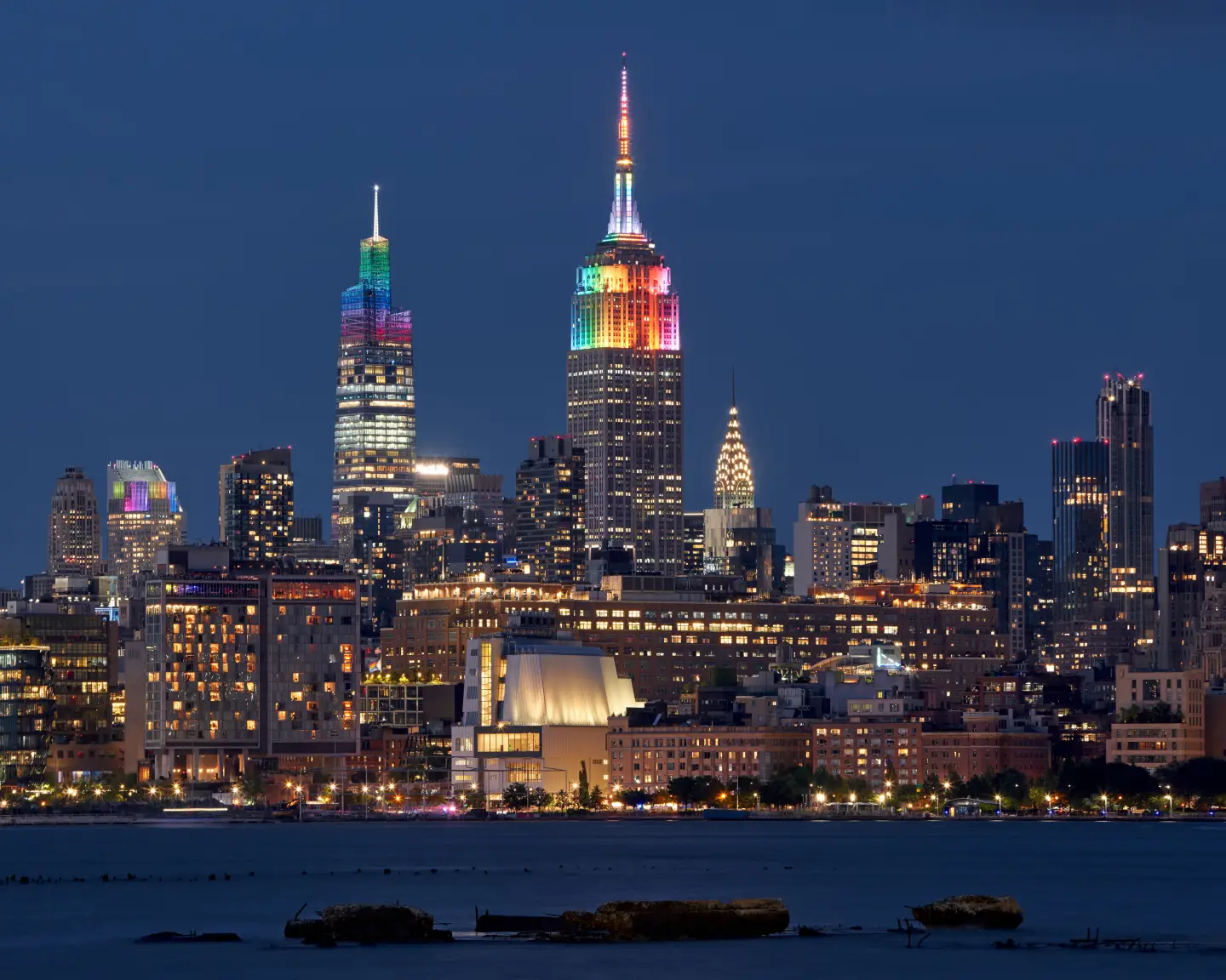 Nächtliche Skyline von Manhattan mit dem bunt beleuchteten Empire State Building im Zentrum, gesehen über den Hudson River
