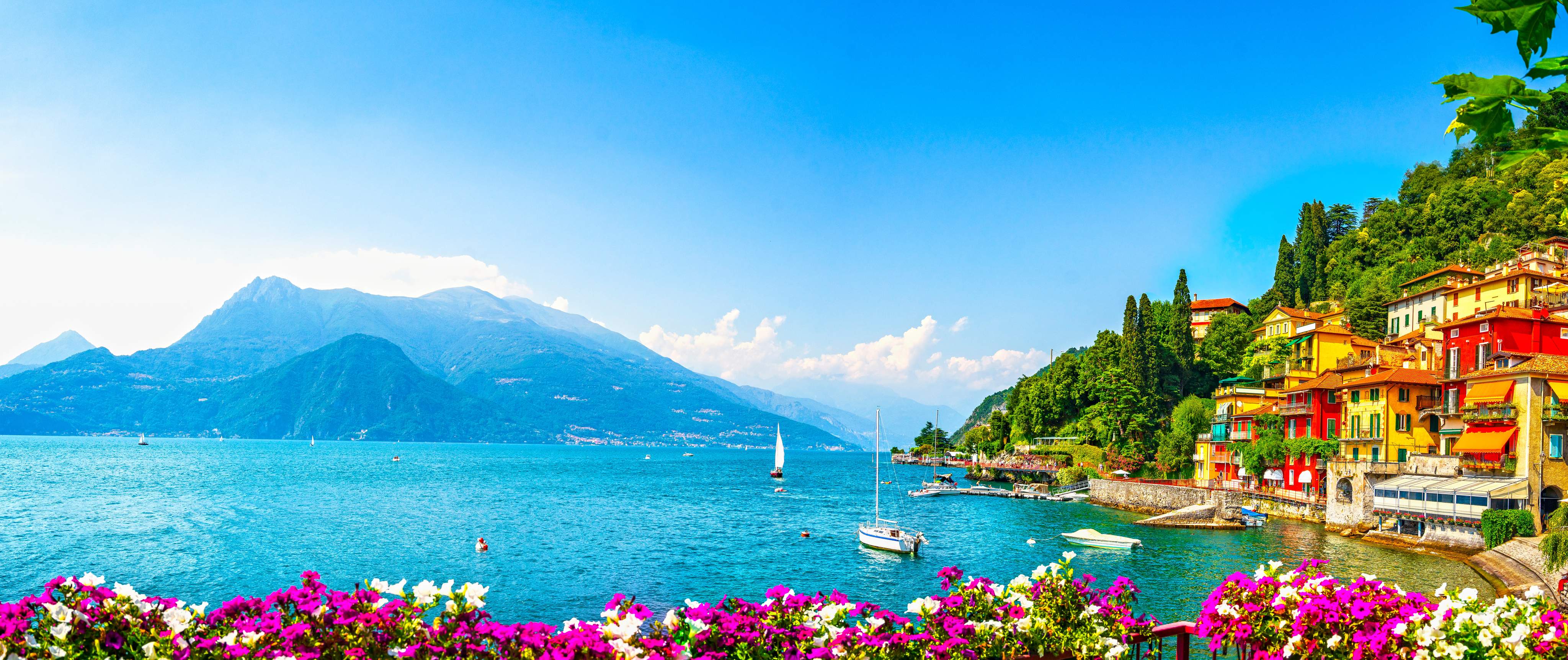 Panoramic view of Varenna on Lake Como with colorful houses on the shore, sailing boats, mountains and blooming flowers in northern Italy.