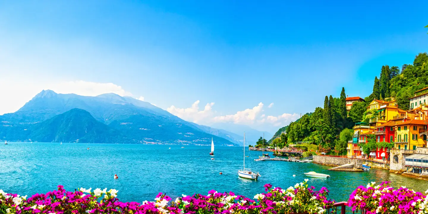 Panoramic view of Varenna on Lake Como with colorful houses on the shore, sailing boats, mountains and blooming flowers in northern Italy.
