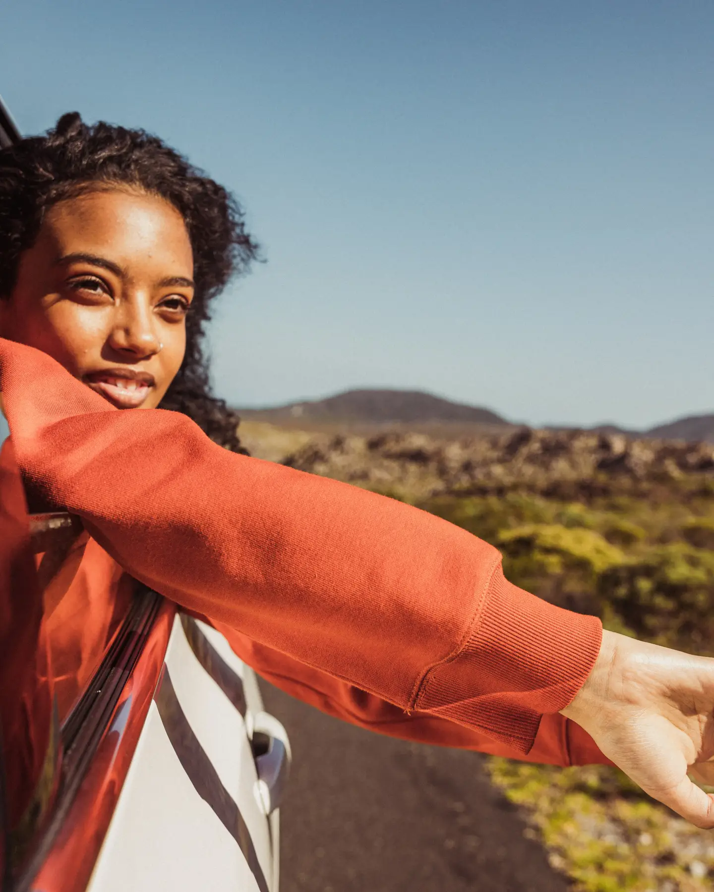 A woman looks out of the window of a car, her arms hanging casually over the door; in the background you can see a desert landscape.
