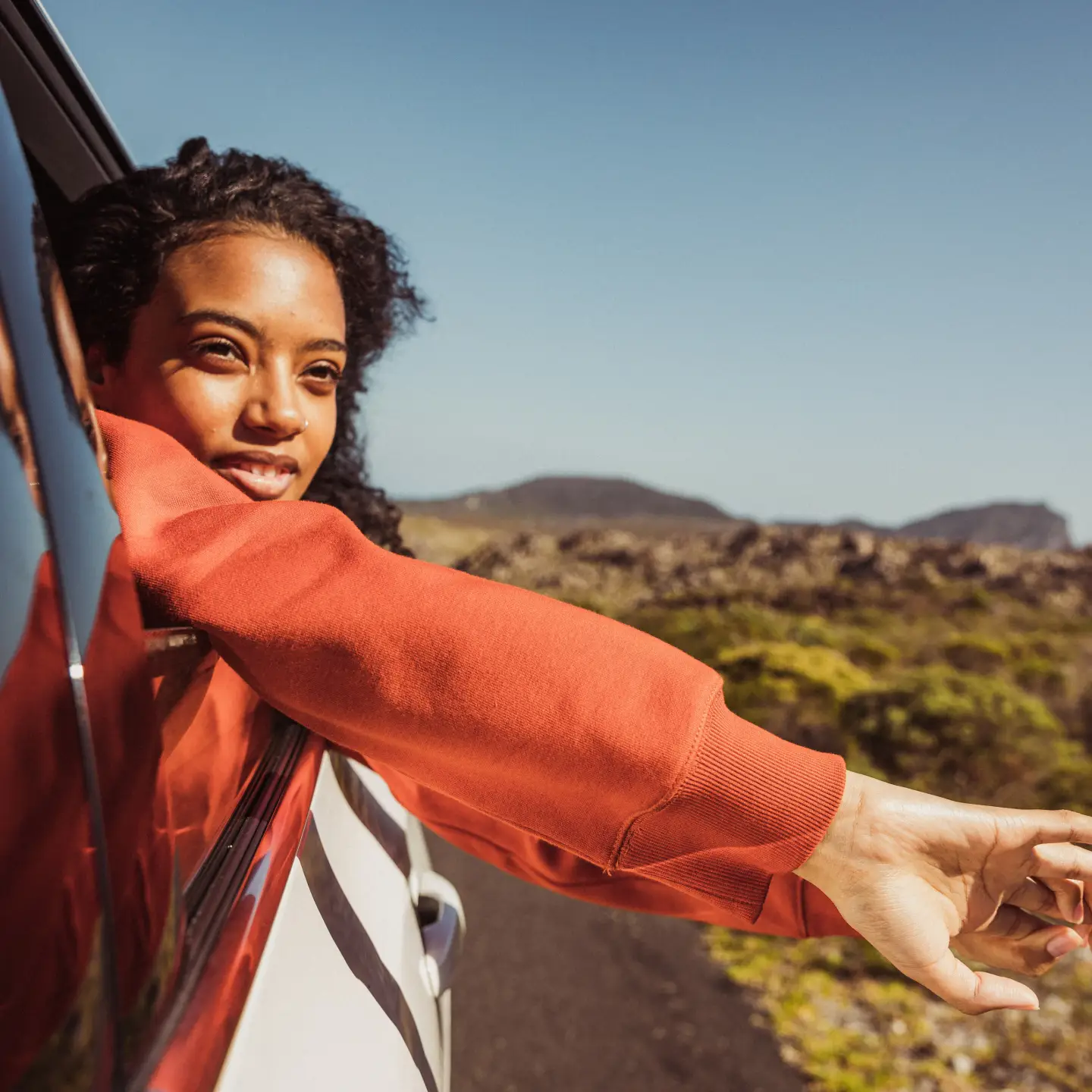 A woman looks out of the window of a car, her arms hanging casually over the door; in the background you can see a desert landscape.