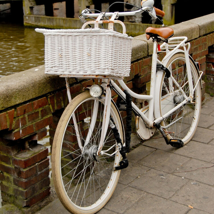 White vintage bicycle with a large wicker basket, parked beside a brick canal wall in Amsterdam