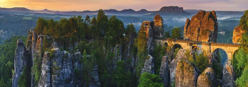 Saxon Switzerland National Park, Germany Dramatic rock formations tower above a lush forest in the Saxon Switzerland National Park, in Germany, while ancient structures crown the horizon under the first light of dawn.