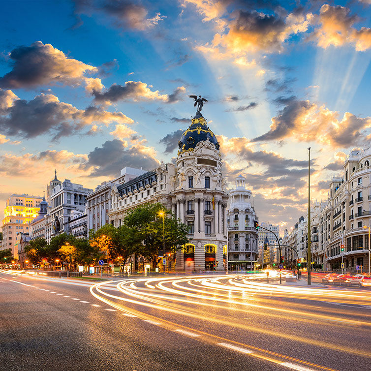 Sunset over a bustling Madrid street, highlighting historic buildings with intricate facades and a majestic statue-topped dome, against a backdrop of vibrant clouds