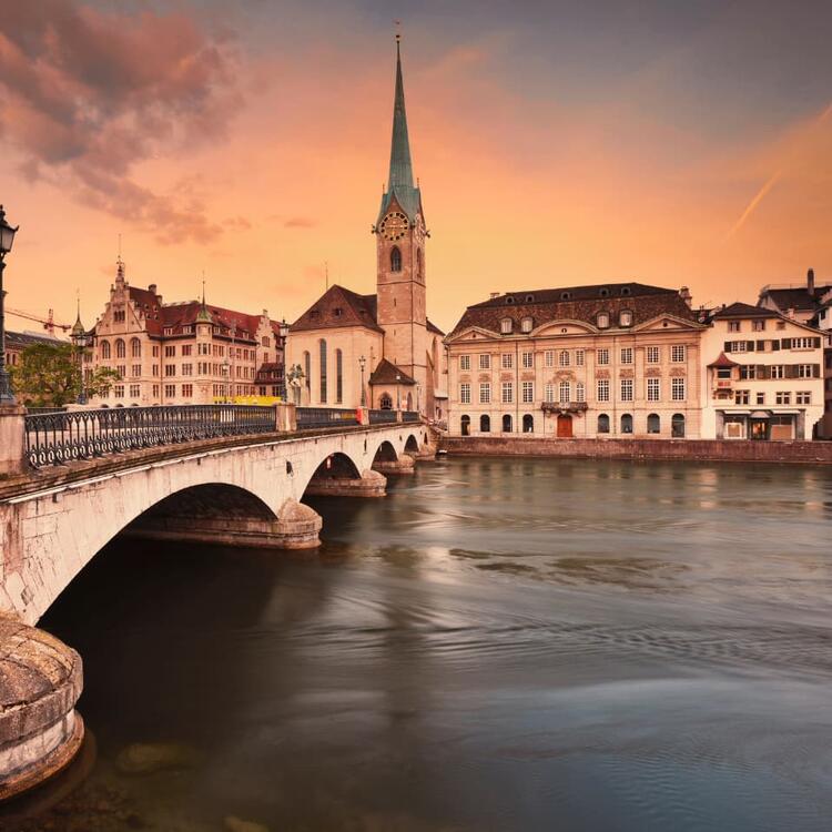 Sunset view of Zurich with the Limmat River in the foreground, an arched stone bridge leading to historic buildings, and the Fraumünster Church with its tall green spire against a backdrop of orange and pink clouds
