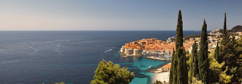 Stunning coastal view of Dubrovnik. The historic town is nestled by turquoise waters, with cypress trees in the foreground and an expansive horizon over the azure sea.