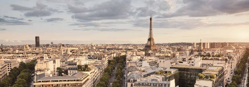 Flights to Paris (CDG) Panoramic view of Paris skyline with the Eiffel Tower standing prominently amidst classic Parisian buildings, under a cloud-streaked sky.