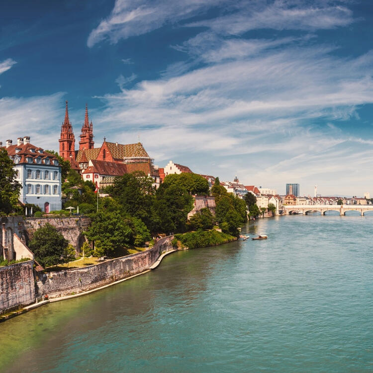Scenic view of Basel in Switzerland with a prominent Gothic cathedral, ornate buildings, and stone walls, overlooking a turquoise river with a bridge, under a blue sky with wispy clouds.