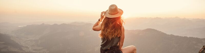 A woman stands with at the top of a mountain, enveloped by the warm hues of sunrise or sunset, embodying tranquility and vastness.