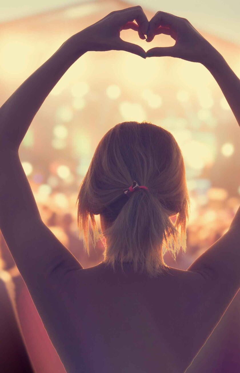 A woman is standing in a festival crowd, holding up her arms and shaping a heart with her hands